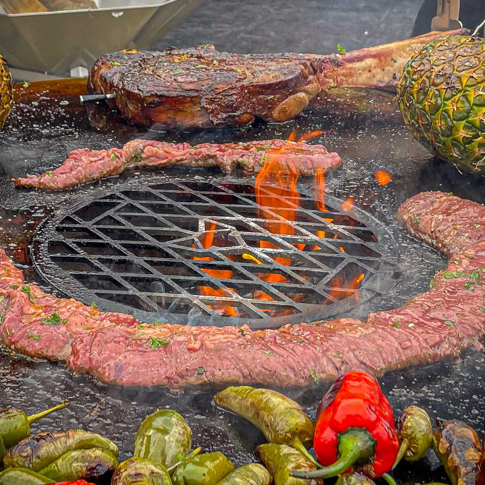 Feuerplatte mit Grillrost im Einsatz. Fleisch und Gemüse und zwei Ananas auf der Feuerplatte.