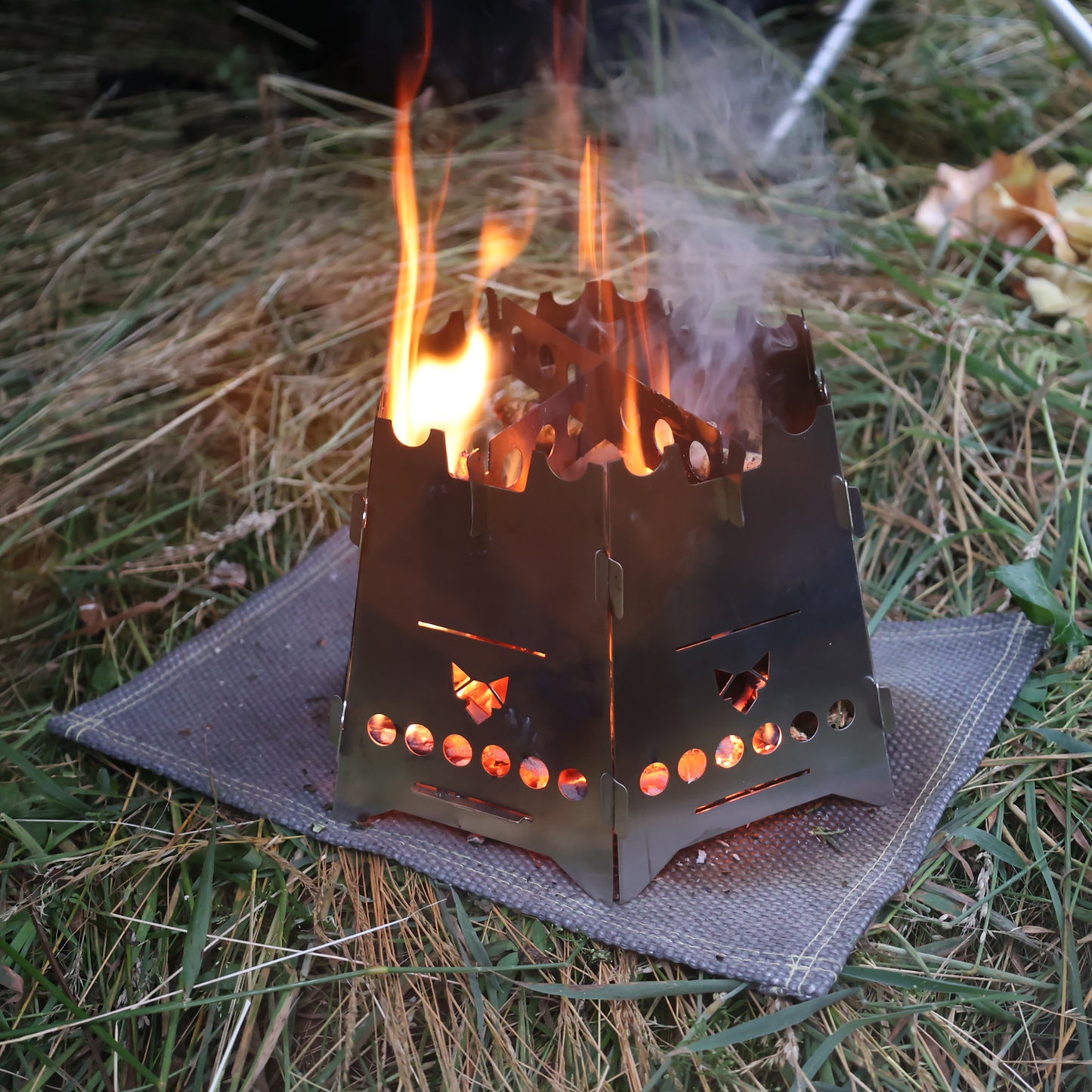 Der Hobokocher FENNEK Basic mit Erweiterungsset mit Flammen darin auf einer Hitzeschutzmatte im Gras
