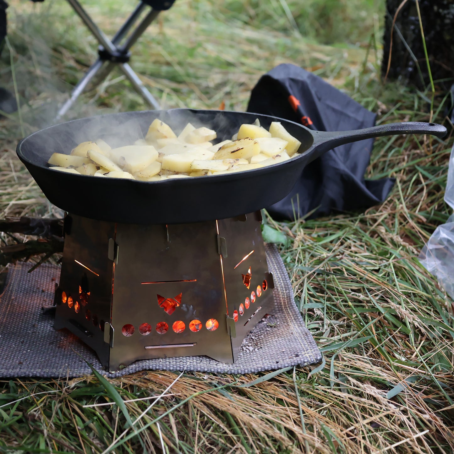 Der Hobokocher FENNEK Basic mit Erweiterungsset in Aktion beim Zubereiten von Bratkartoffeln mit einer schwarzen Gußpfanne auf einer Wiese.