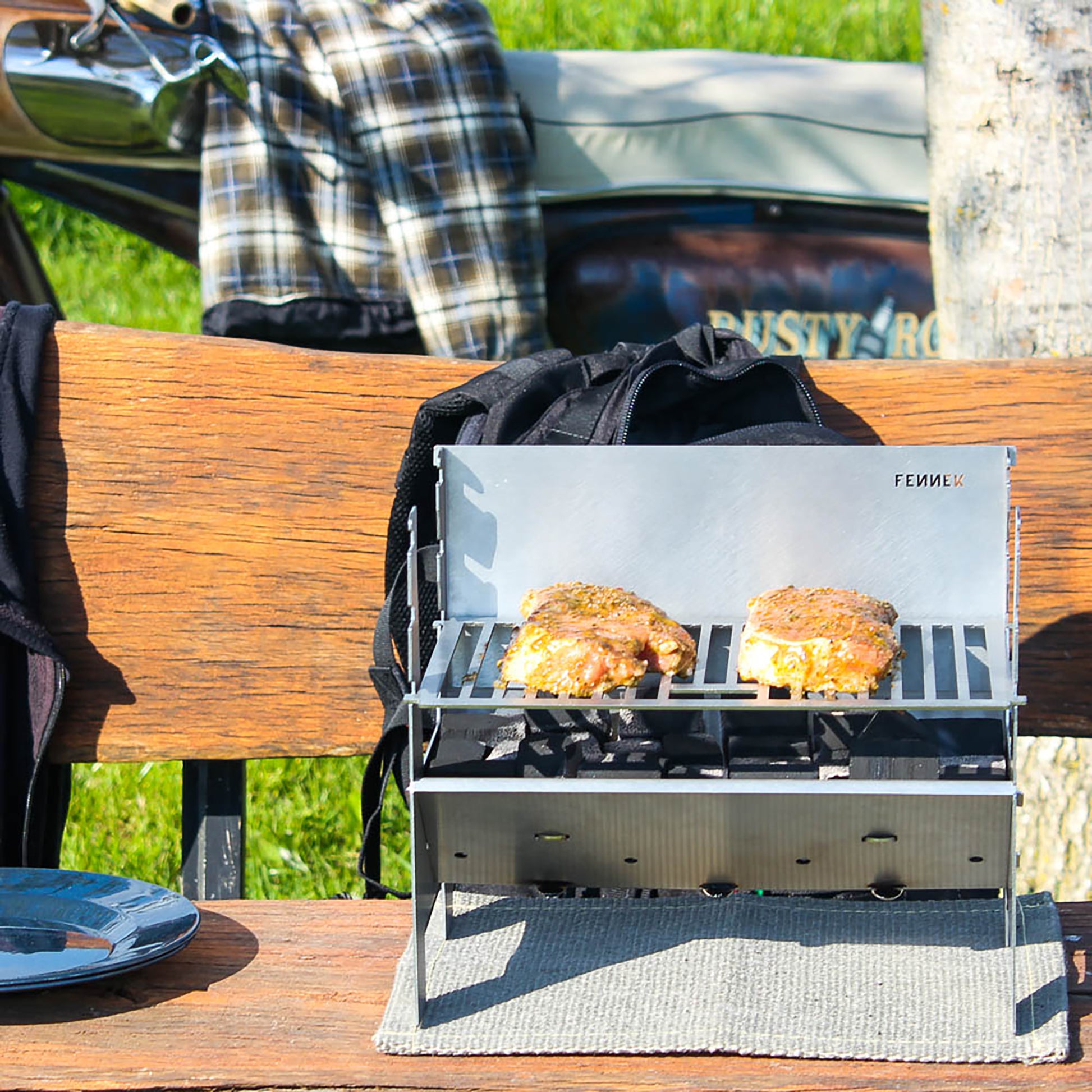 FENNEK Light Holzkohlegrill steckbar, faltbar und mobil, klein und kompakt, steht aufgebaut auf einer Hitzeschutzmatte auf einer Bank. Es werden Steaks gegrillt.
