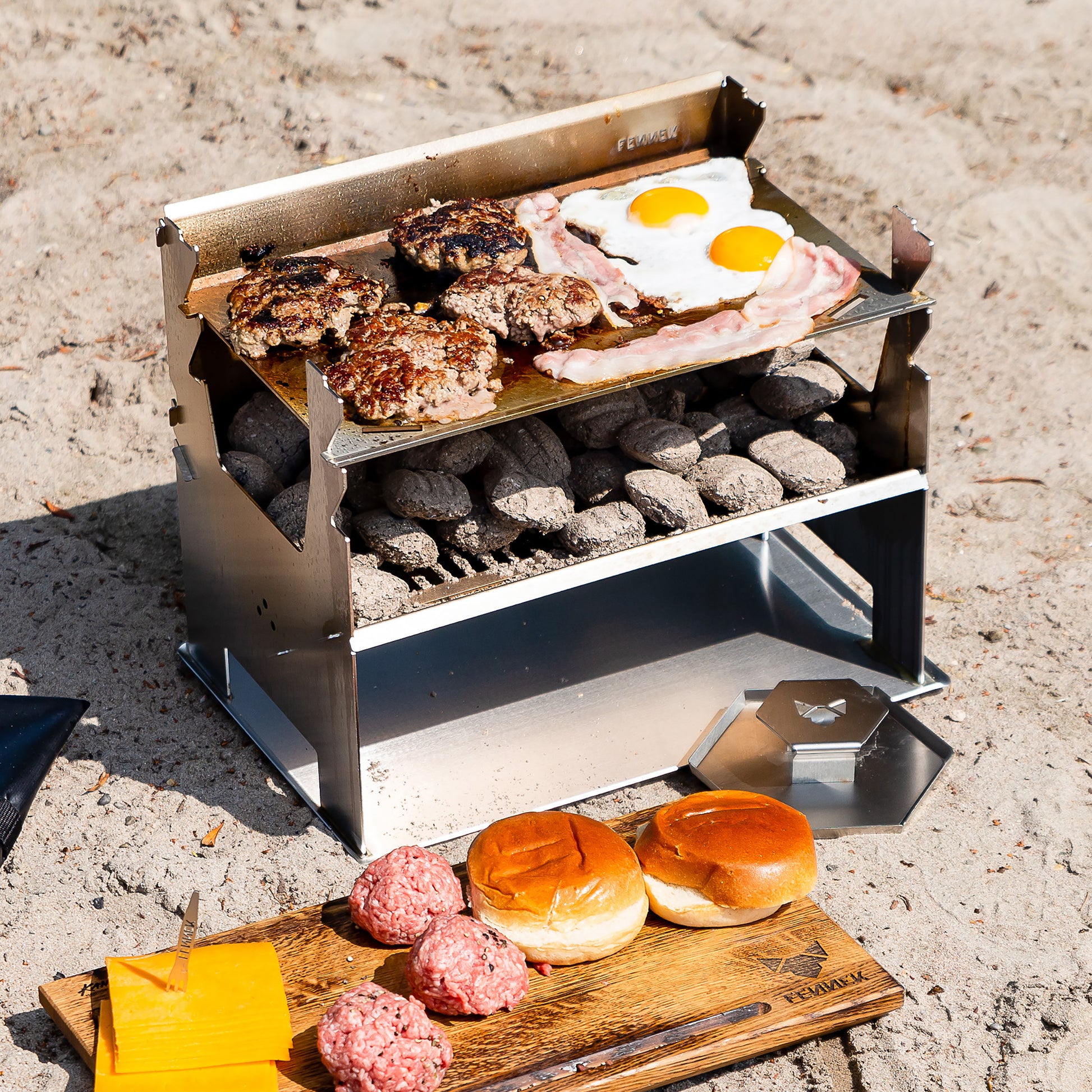 Edelstahlholzkohlegrill mit Briketts grillt Burger und Spiegelei auf einer Plancha und steht auf einem Bodenblech im Sand, davor Brötchen und eine Edelstahl Burgerpresse