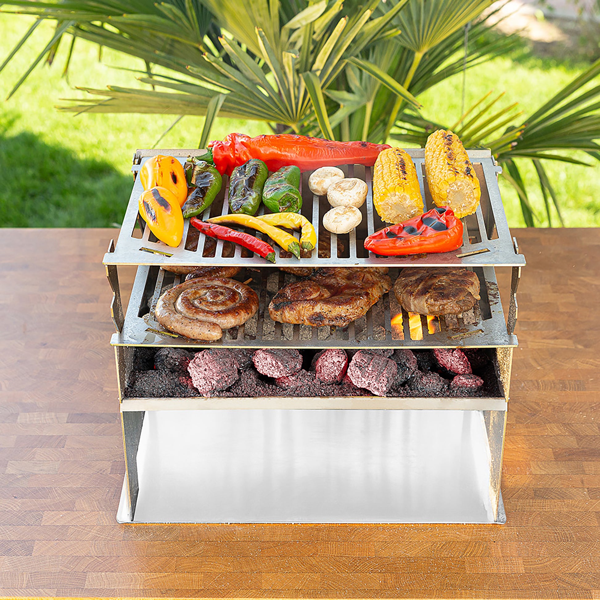 Holzkohlegrill aus Edelstahl mit 2 übereinander platzierte Grillroste und reichlich Fleisch und buntem Gemüse darauf.