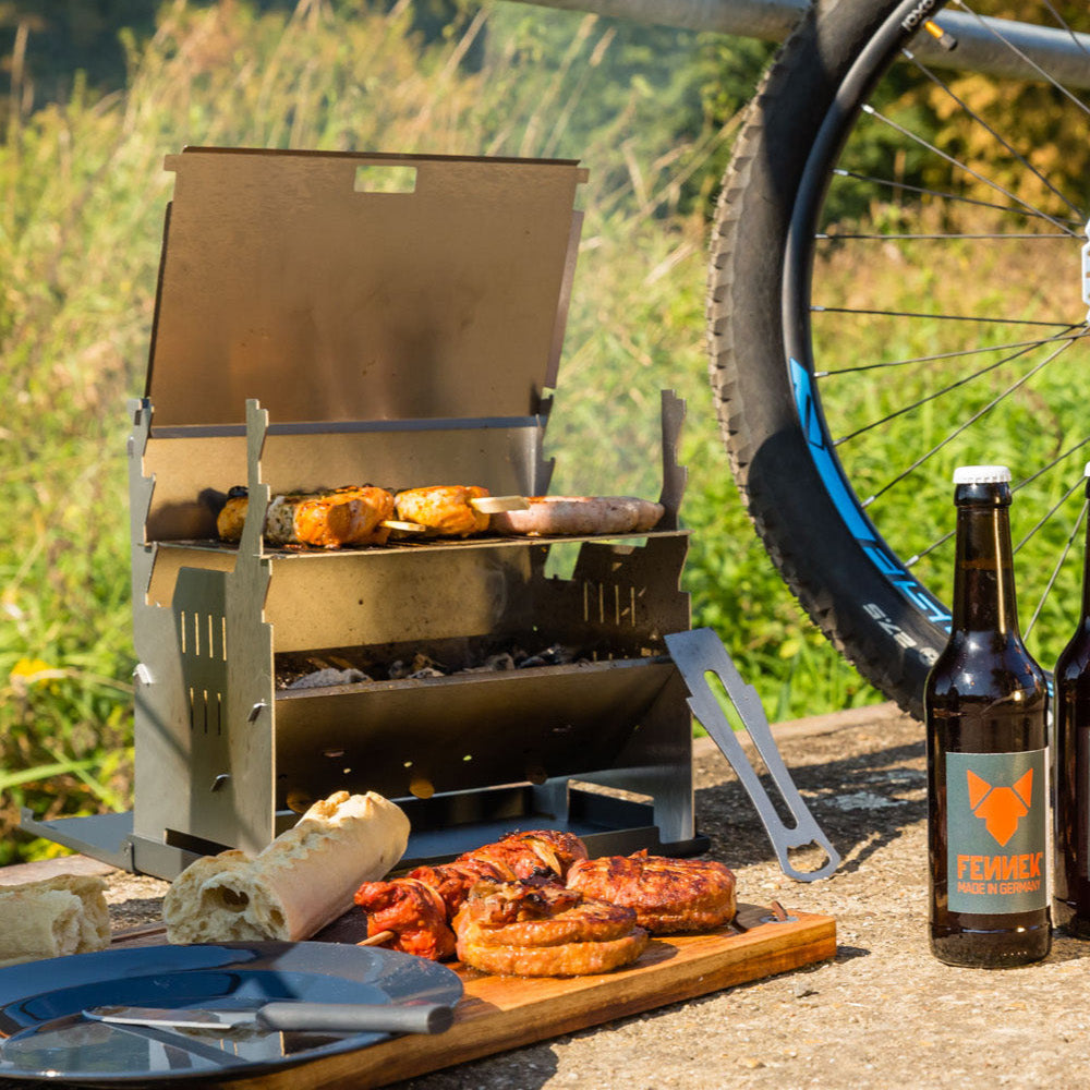 Der Grill steht neben einer Wiese auf dem Boden. Auf dem Grill brutzeln Spieße und Würstchen. Davor liegt das FENNEK Schneidebrett mit Teller, Besteck, Baguette und gegrilltem Fleisch. Daneben stehen zwei Flaschen und im Hintergrund steht ein Fahrrad.