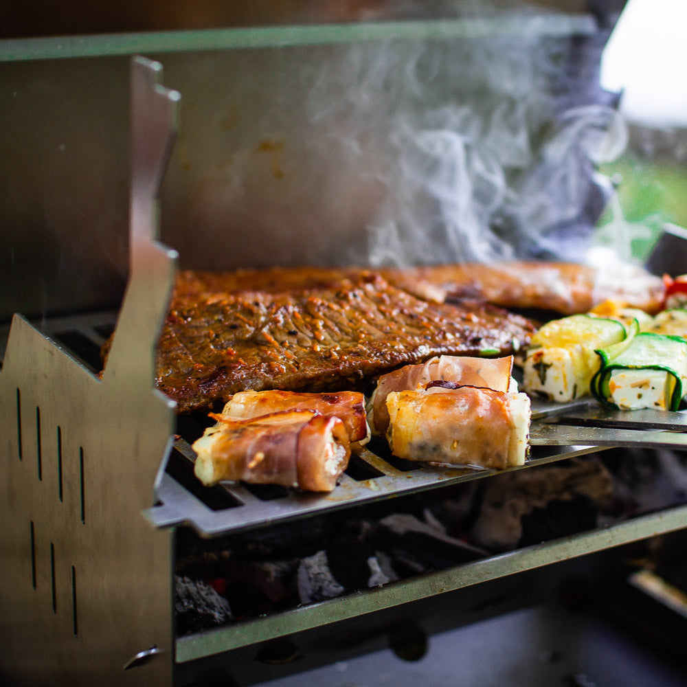 Bild zeigt den FENNEK Grill in Betrieb. Auf dem Grillrost brutzelt unter anderem Gemüse und Fleisch vor sich hin. Außerdem ist der dazugehörige Rostheber eingehakt. Dieser kann aber auch als Flaschenöffner genutzt werden.