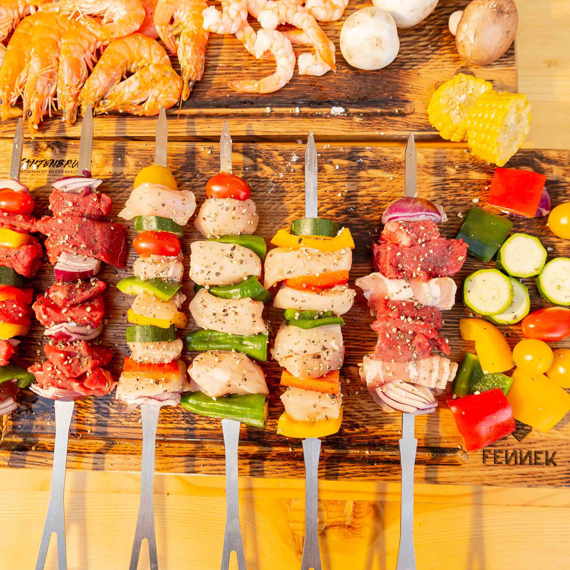 Mehrere kurze Grillspieße zum Grillen vorbereitet, liegen bunt bestückt mit Gemüse und Fleisch oder Geflügel auf einem Holzbrett.