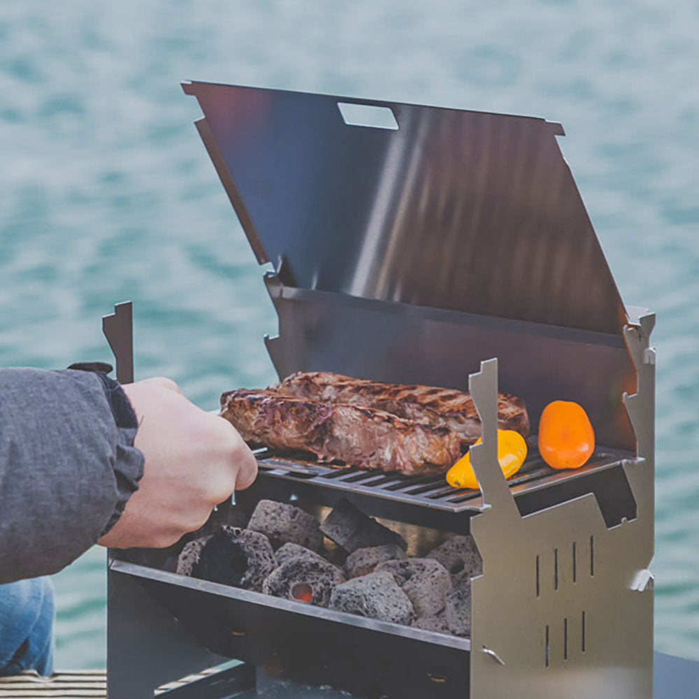 Bild zeigt den FENNEK Grill am See im Action. Ein Mann grillt sein Fleisch und Gemüse.