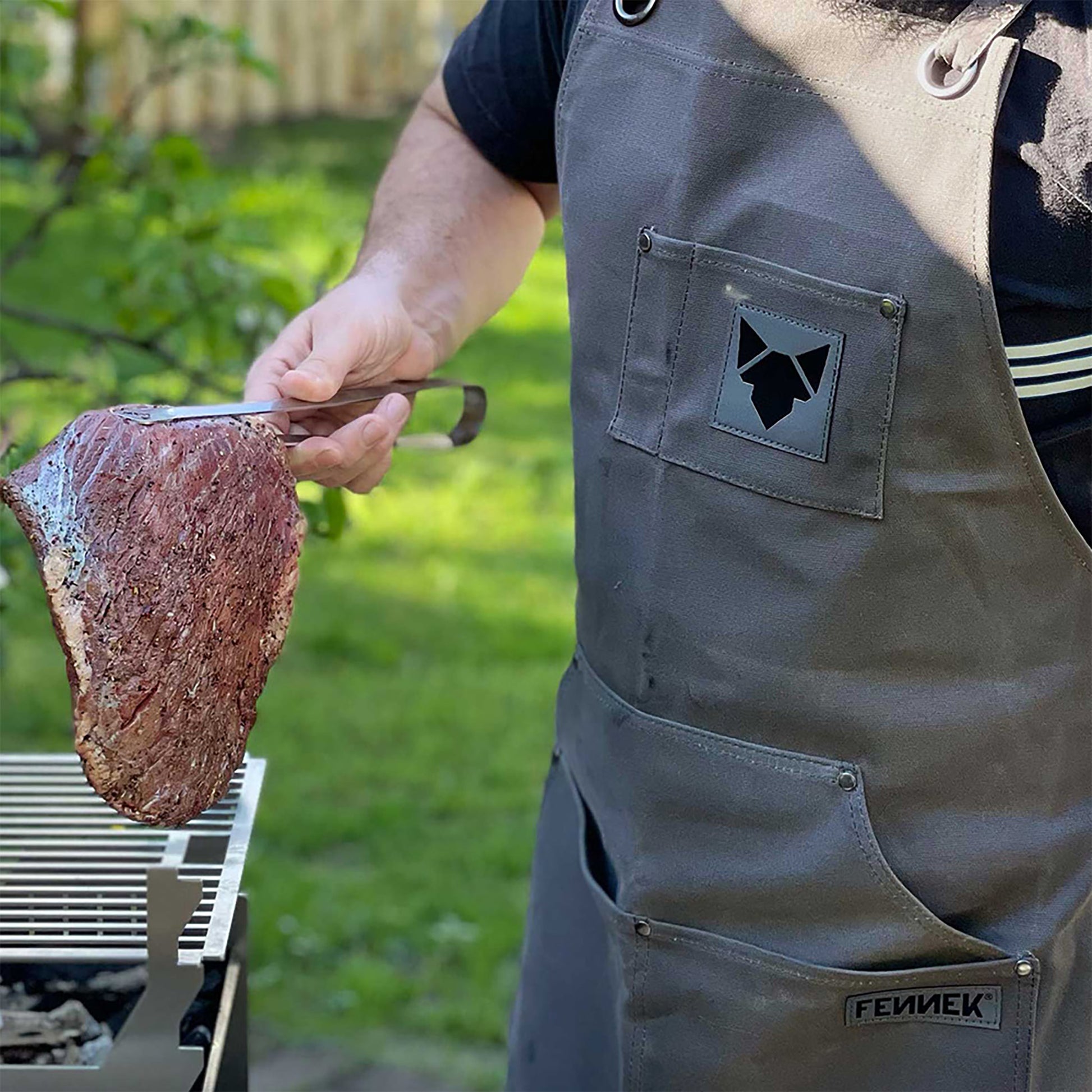 Griller mit FENNEK Grillschürze hält in der Hand mit einer Grillzange ein große Stück mariniertes Grillfleisch im heimischen Garten.