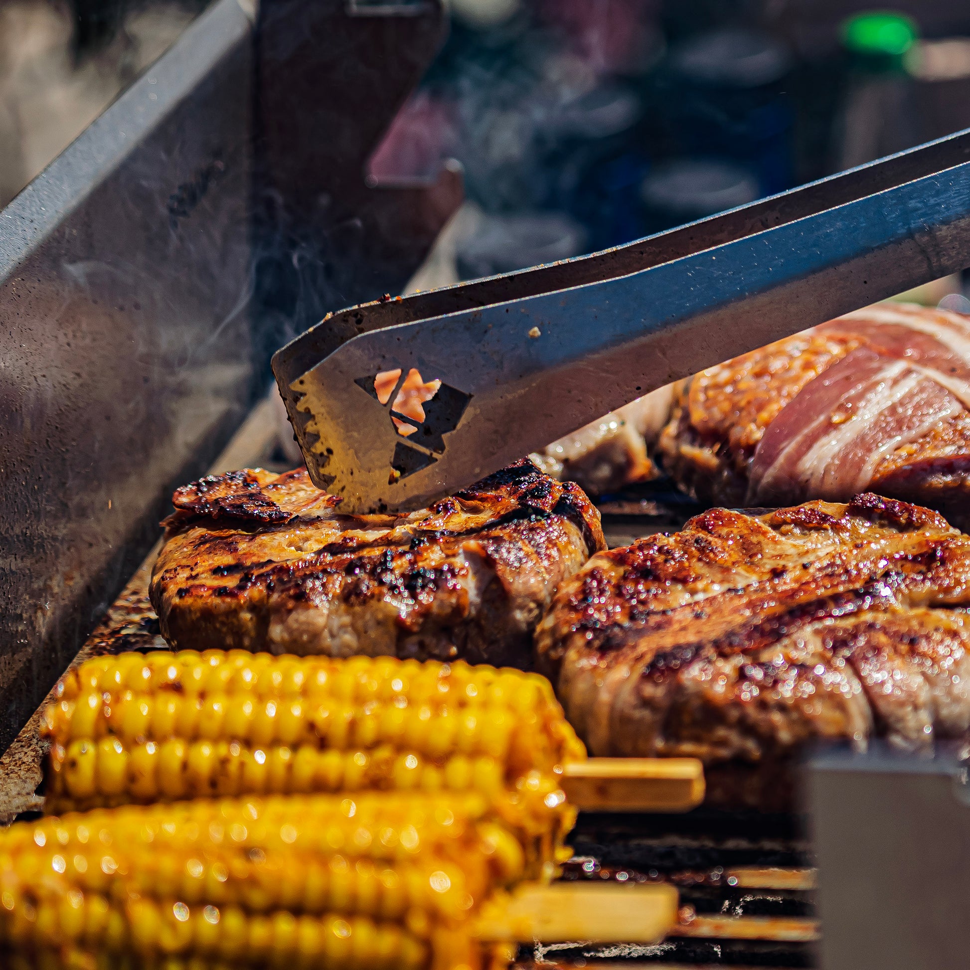 Auf dem Grill brutzeln Fleisch und Maiskolben, welche mit der FENNEK Grillzange gewendet werden.