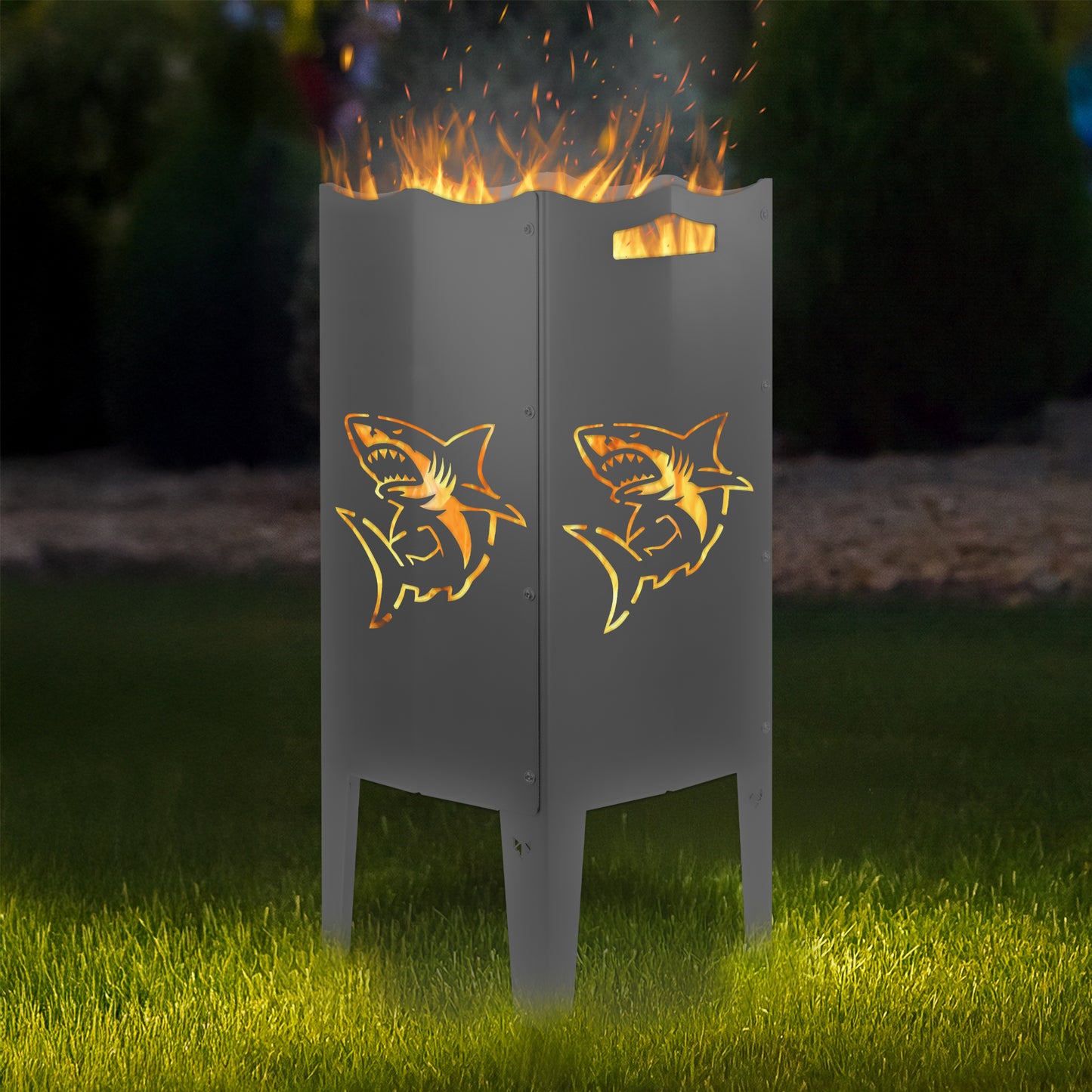 FENNEK Feuersäule Stahl mit dem Motiv Hai - in der Feuersäule brennt ein Feuer, man sieht das Motiv sehr schön leuchten in der Abenddämmerung in einem Garten. Die Feuersäule steht auf Gras.