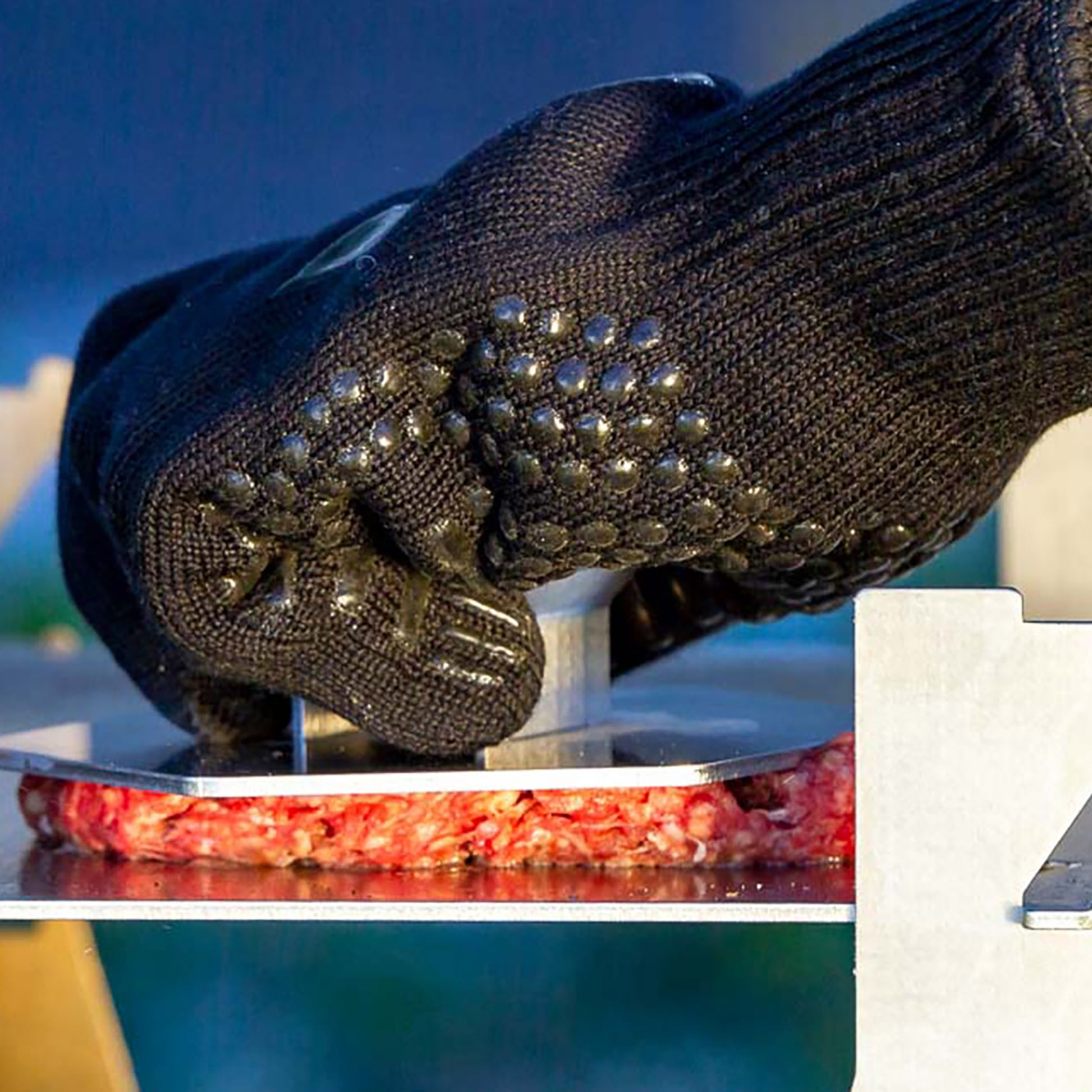 Nahaufnahme wie ein schwarzer Grillhandschuh mit einer Burgerpresse Hackfleisch auf einer Metallplatte flachpresst