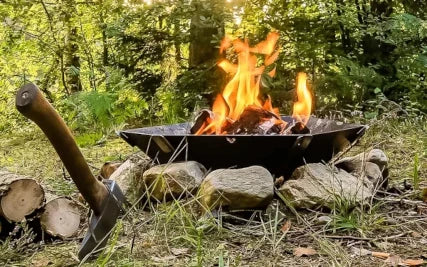 Feuerschale Hexagon im Wald auf Steinkreis. Darin ein Feuer. Vorne links eine Axt im Boden und 2 Feuerholz.