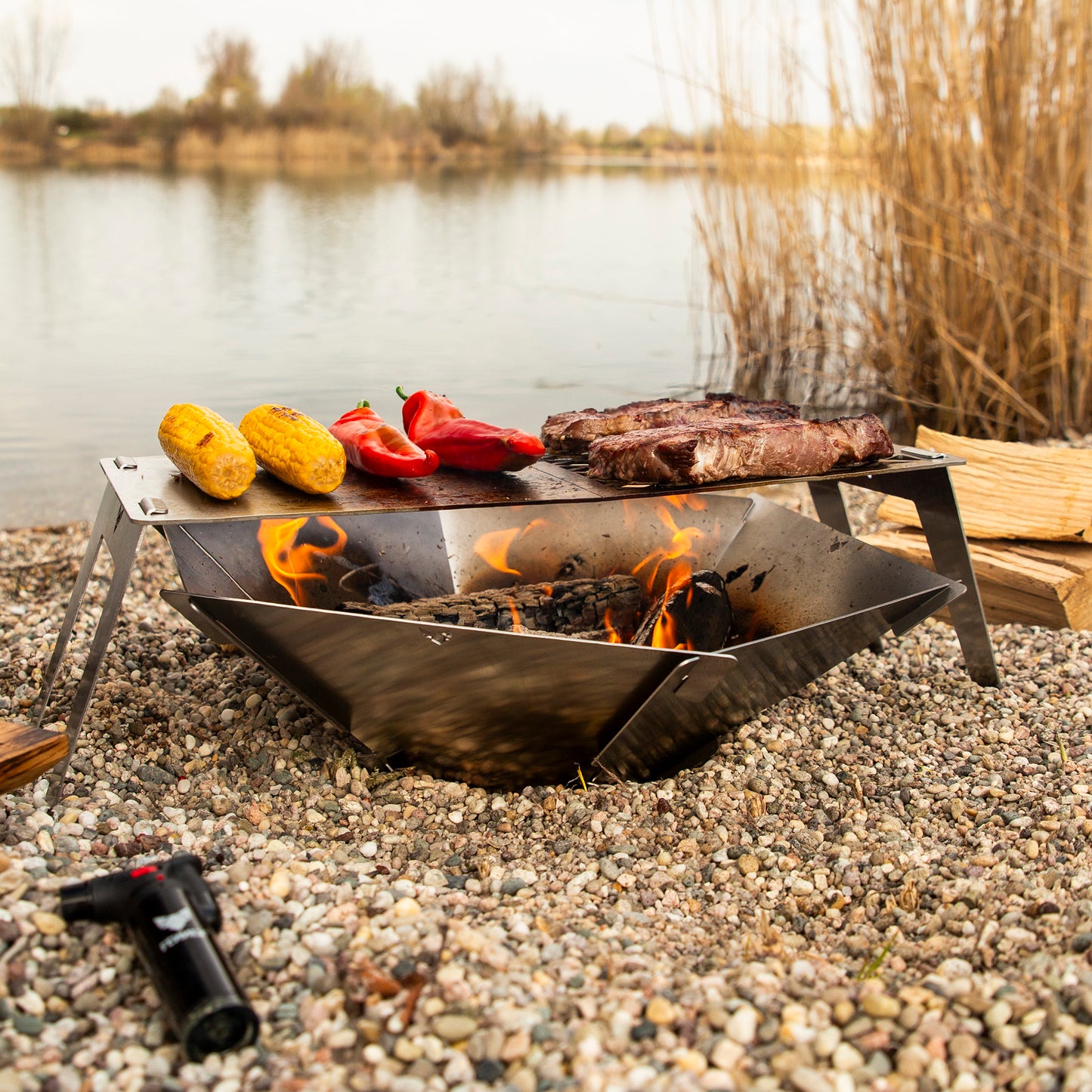 Die FENNEK Feuerschale Hexagon steht in einem Kiesbett am See. Darüber ist das FENNEK 4Fire aufgestellt, auf welchem zwei Steaks und Gemüse gegrillt wird.