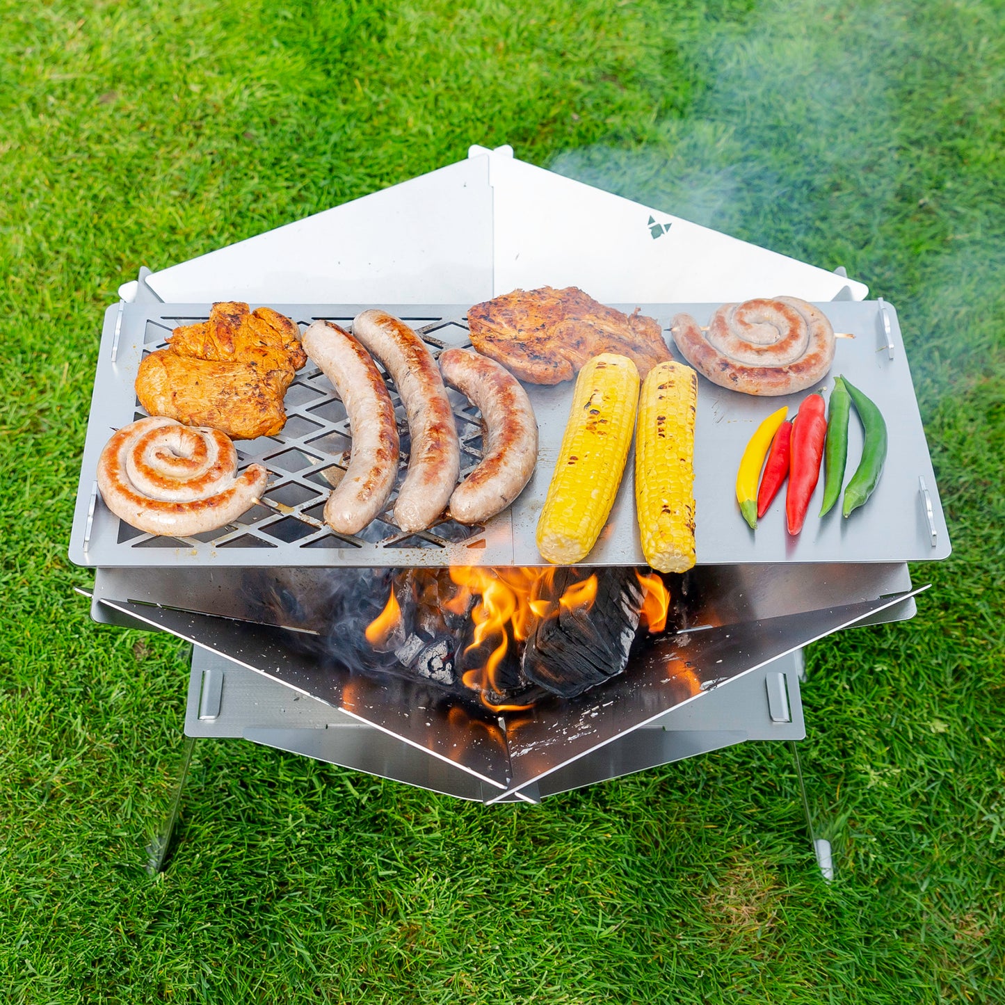 Fleisch und Gemüse wird auf einer zusammengesteckten Grillplatte über einer Feuerschale gegrillt die sich mit Erhöhung auf einem grünen Rasen befindet.