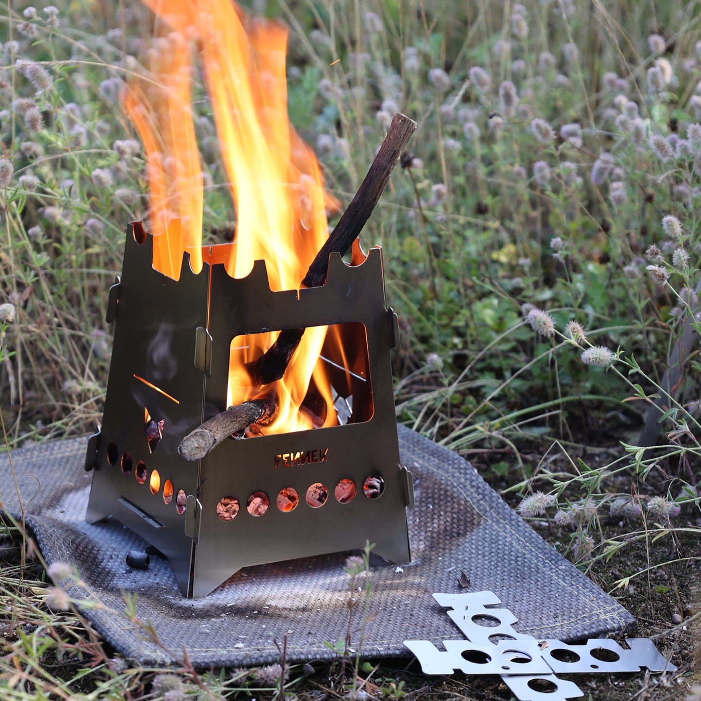 Brennendes Holz im Hobokocher darunter eine Hitzeschutzmatte auf einer Wildwiese.