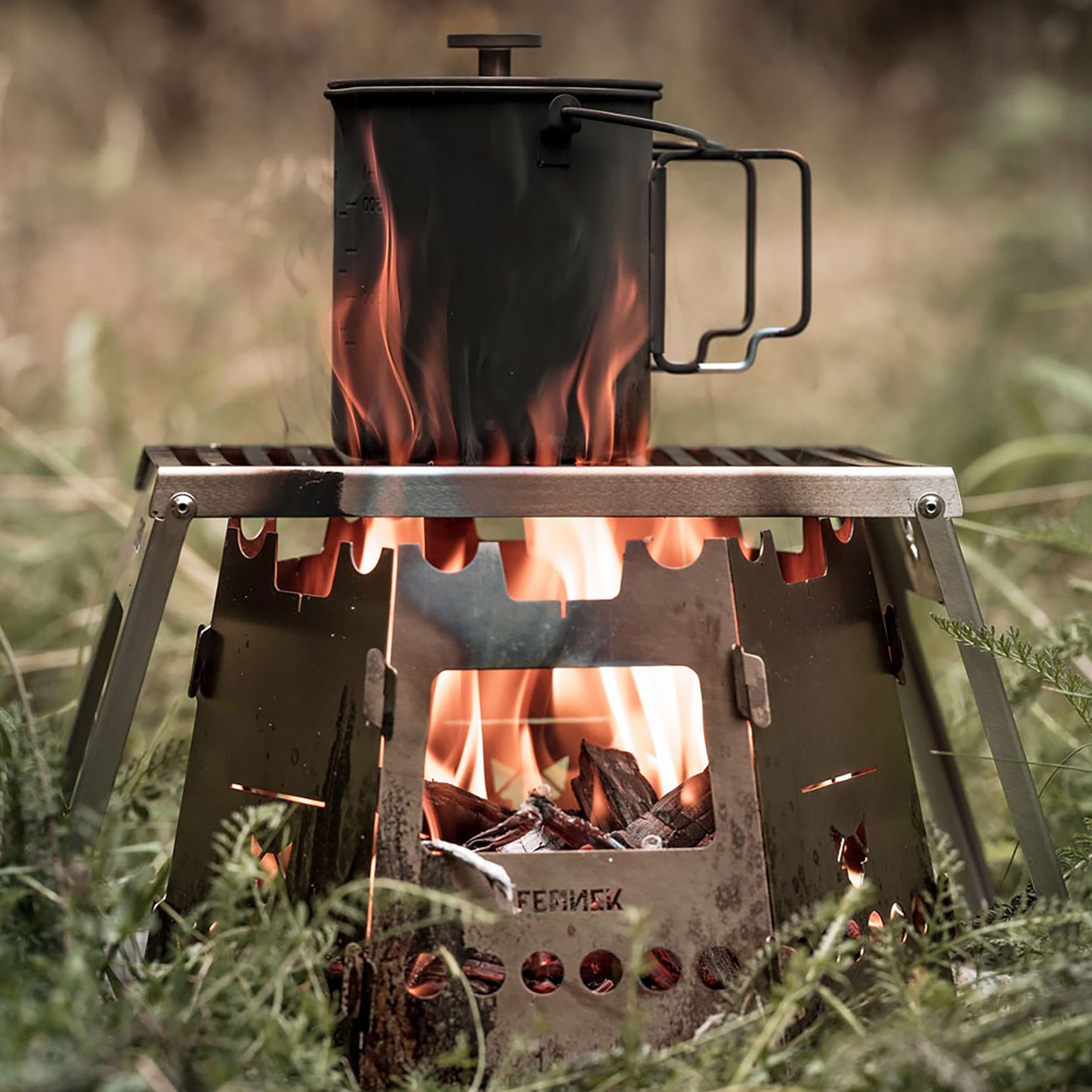 Outdoortasse auf einem Klapprost über einem Feuer das im Hobokocher mit Erweiterungs-Set brennt in der grünen Natur.