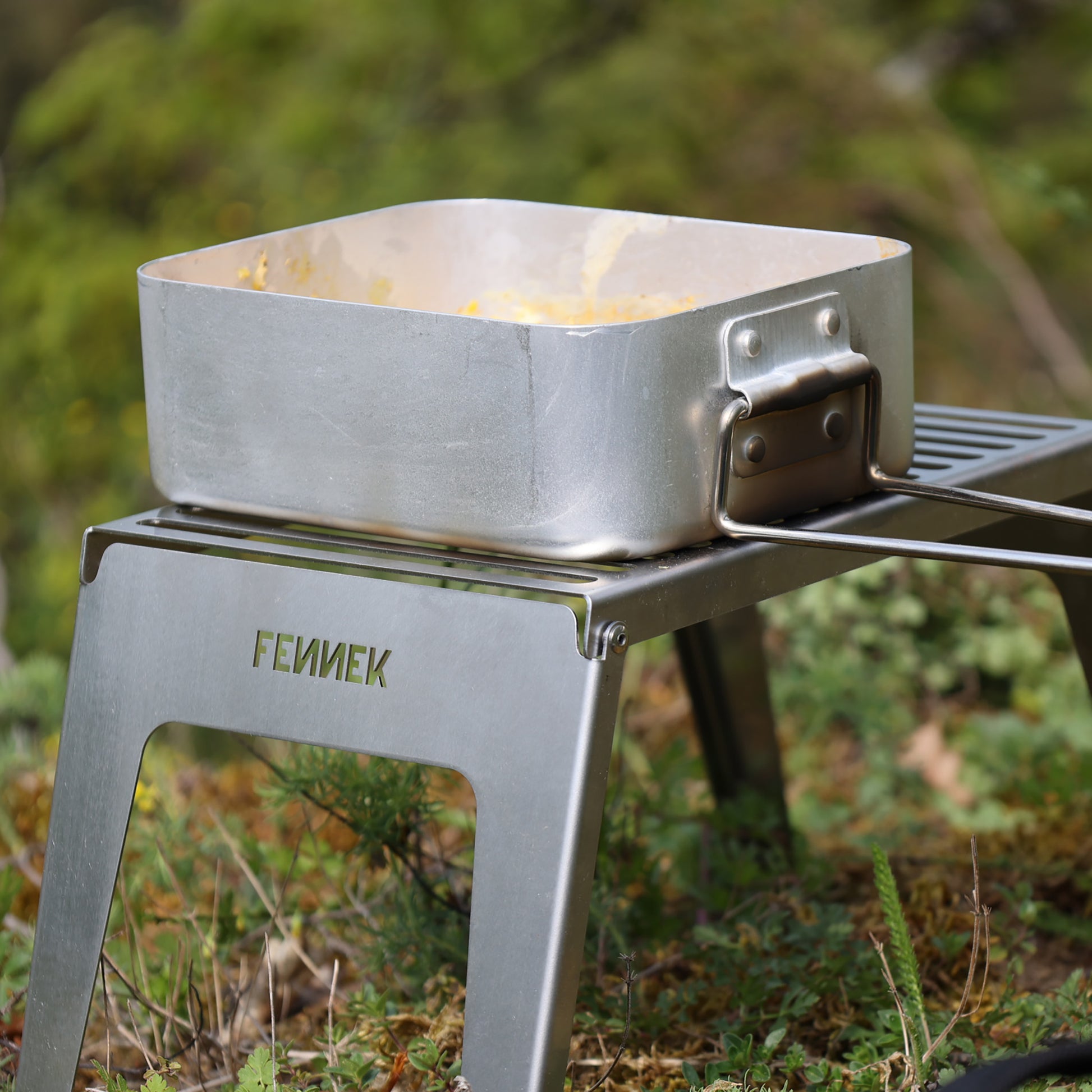 Ein rechteckiges Kochgeschirr mit abgerundeten Ecken und ausgeklappten Griff steht auf dem in der Natur aufgestellten Klapprost.