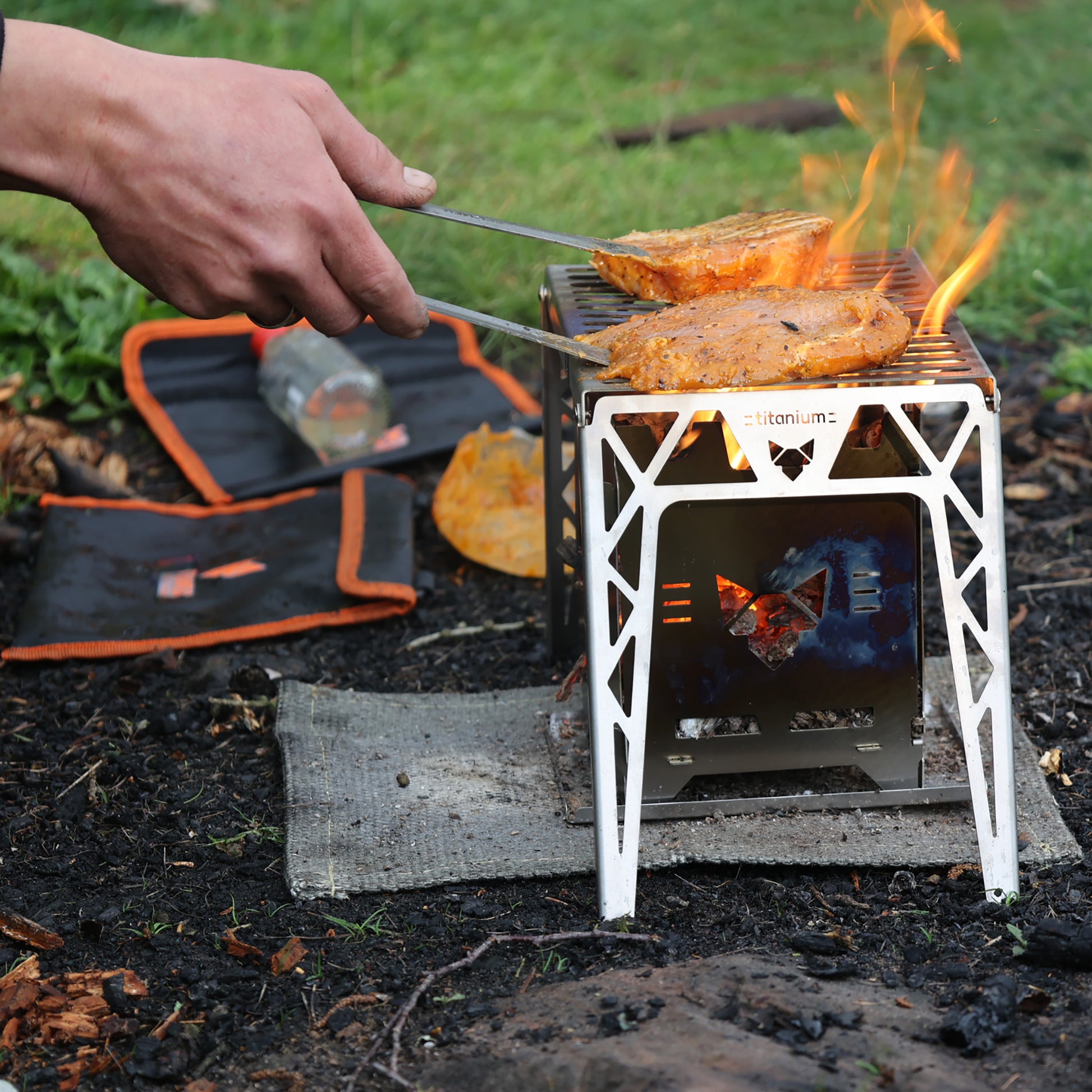 Mariniertes Geflügel-Filet das auf dem Klapprost mit FENNEK Titanium-Schriftzug liegt und in der Natur über den Flammen eines Hobokochers zubereitet wird. Eine Hand mit Grillzange wendet das Fleisch.