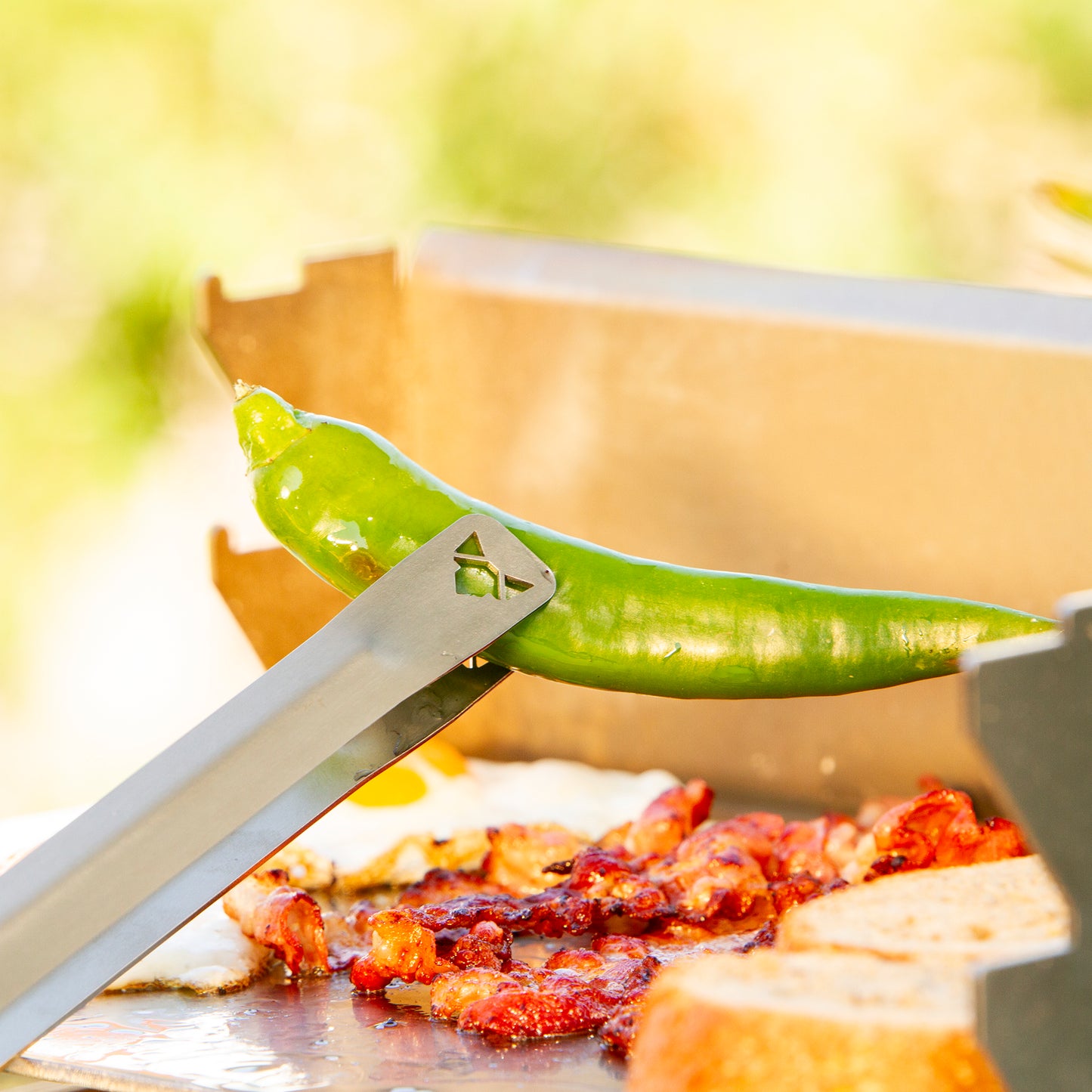 Edelstahl Grillzange mit FENNEK-Logo an der Kontaktfläche herausgeschnitten hält eine Pepperoni von der Plancha.