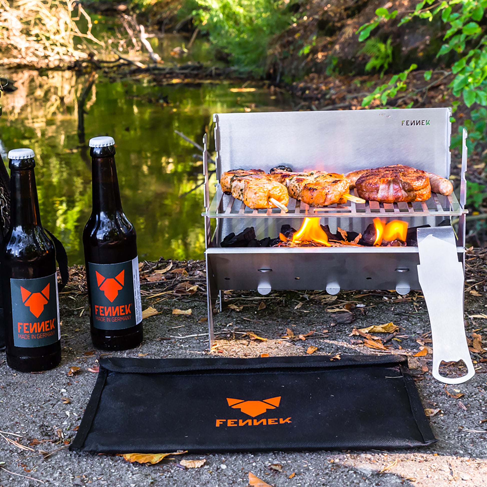 Im Freien aufgebauter Fennek Grill aus Edelstahl auf dem Grillfleisch über brennender Kohle gegrillt wird, daneben 2 Flaschen mit FENNEK-Etikett und vor dem Grill liegt die schwarze Nylon-Tasche mit FENNEK Logo-Aufdruck, unscharf im Hintergrund Gewässer und viel grüne Natur.
