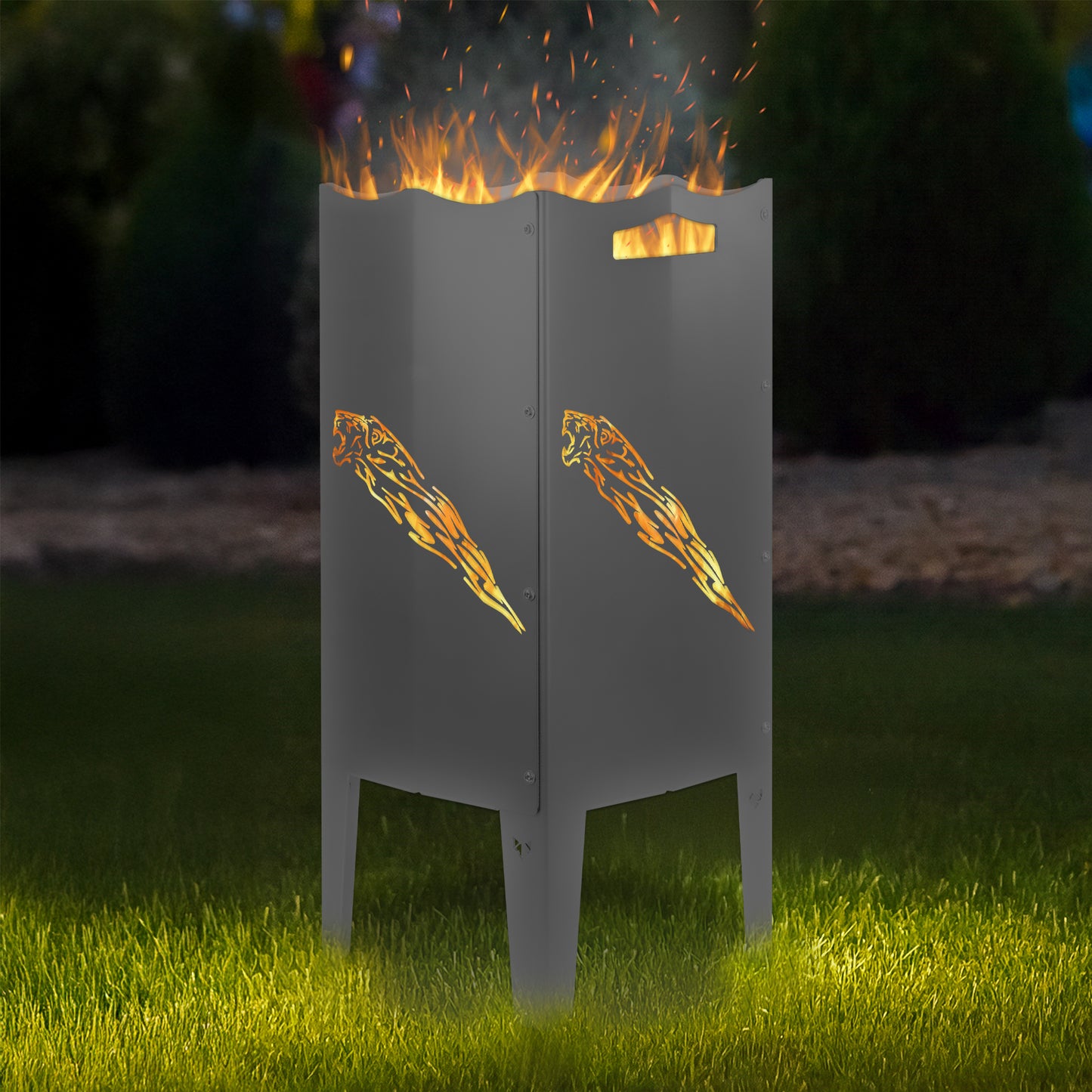 FENNEK Feuersäule Stahl mit dem Motiv Löwe mit Schweif - in der Feuersäule brennt ein Feuer, man sieht das Motiv sehr schön leuchten in der Abenddämmerung in einem Garten. Die Feuersäule steht auf Gras.