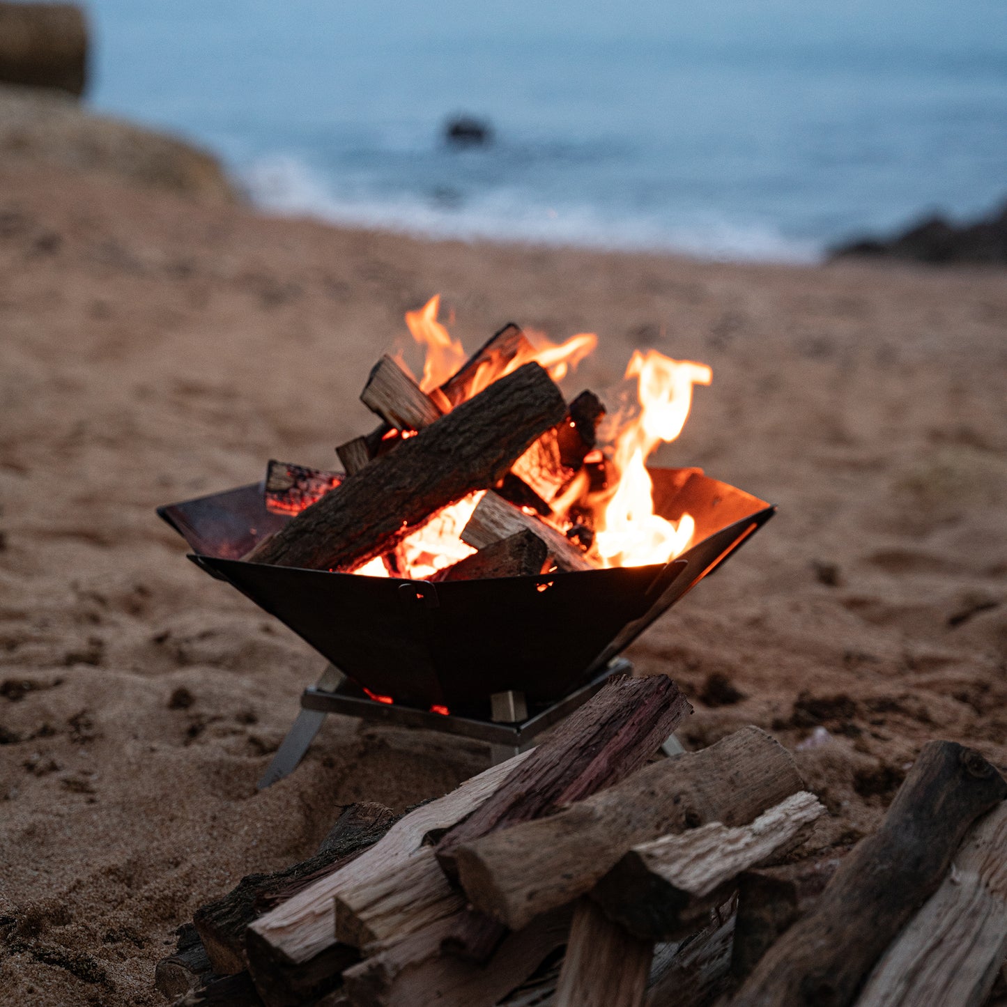 Holz brennt in einer mobilen und steckbaren Edelstahl Feuerschale steht mit 4 Standfüßen auf einem Strand.