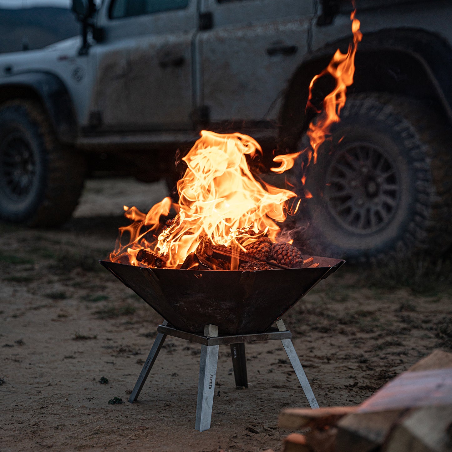 Die FENNEK Feuerschale Oktagon mit den optionalen Füßen steht vor einem Land Rover am Strand. Es ist Abendstimmung. Ein schönes Feuer lodert in der Feuerschale.