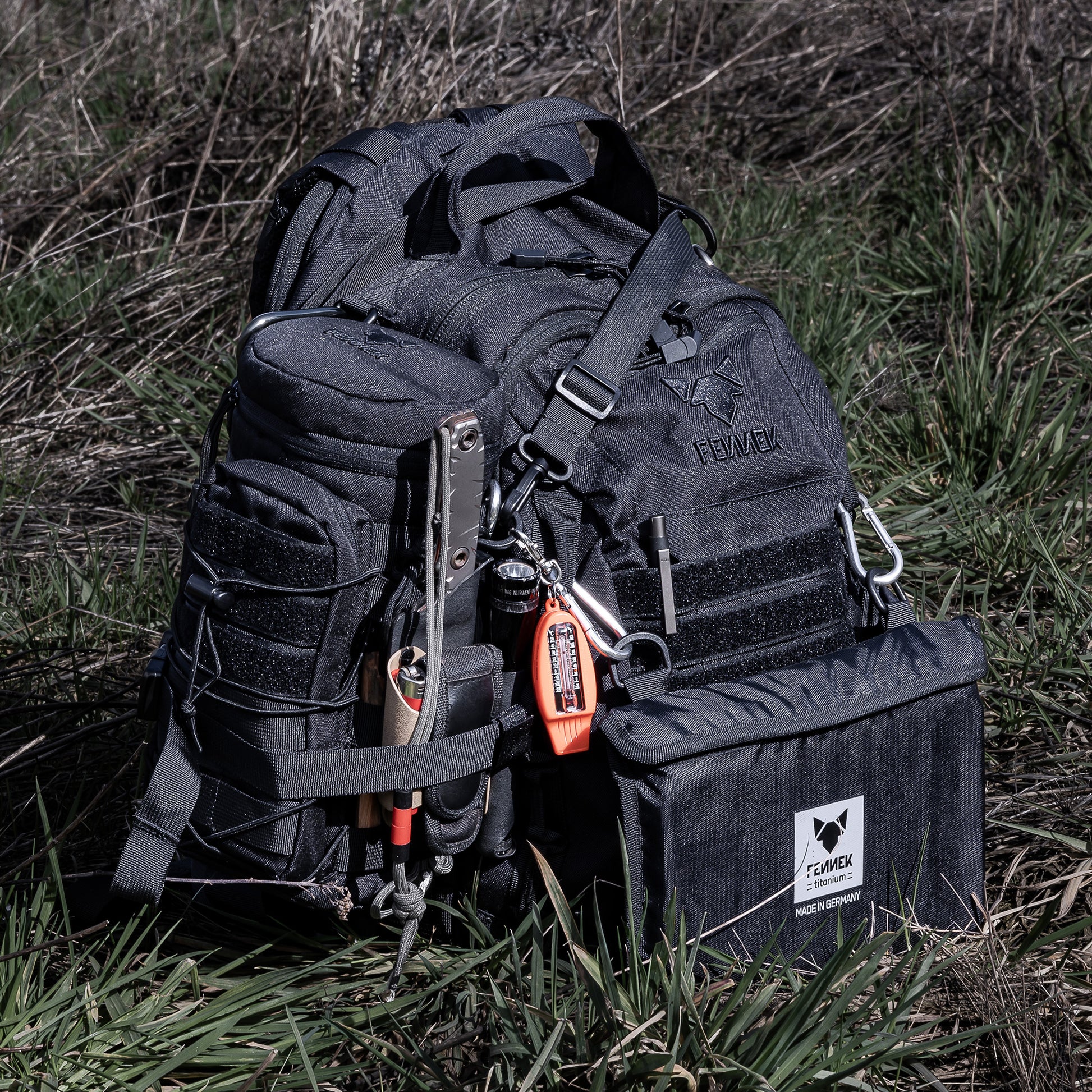 Der Backpack One2explore steht auf einer Wiese. Die Zubehörtasche ist seitlich mit den Kompressionsgurten am Backpack festgeschnallt. An der Vorderseite befindet sich der  Klapprost Titan in seiner dazugehörigen Tasche, mithilfe von Karabinern ist dieser am Backpack befestigt.