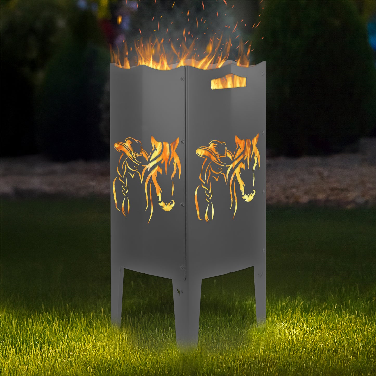 FENNEK Feuersäule Stahl mit dem Motiv Pferd & Frau - in der Feuersäule brennt ein Feuer, man sieht das Motiv sehr schön leuchten in der Abenddämmerung in einem Garten. Die Feuersäule steht auf Gras.