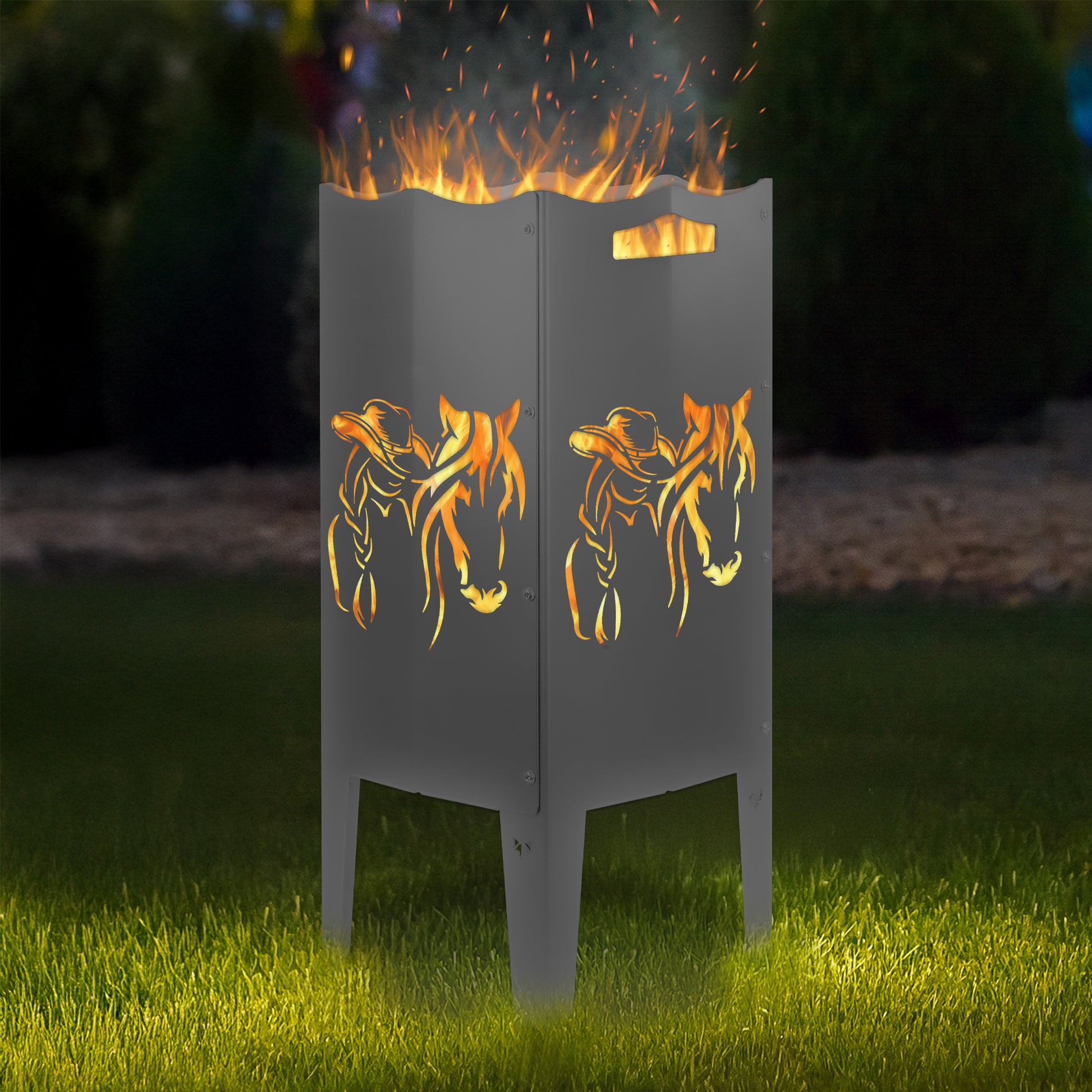 FENNEK Feuersäule Stahl mit dem Motiv Pferd & Frau - in der Feuersäule brennt ein Feuer, man sieht das Motiv sehr schön leuchten in der Abenddämmerung in einem Garten. Die Feuersäule steht auf Gras.