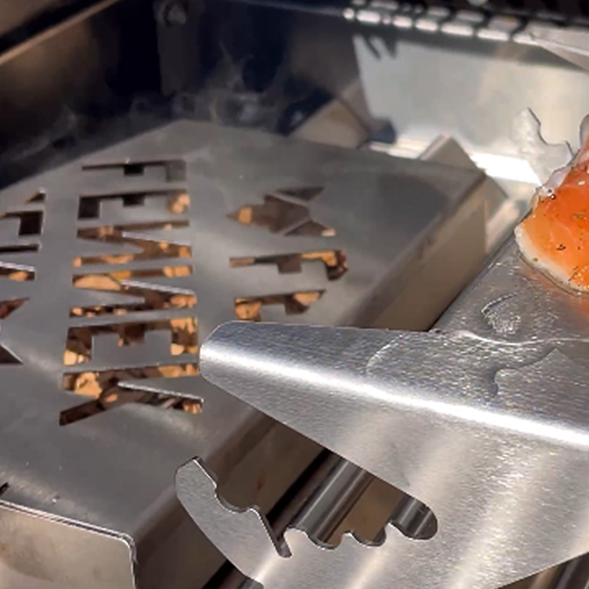 Der Bildausschnitt zeigt den FENNEK Flammlachshalter mit Lachs auf dem Grillrost von einem Gasgrill. Daneben befindet sich die FENNEK Räucherbox.