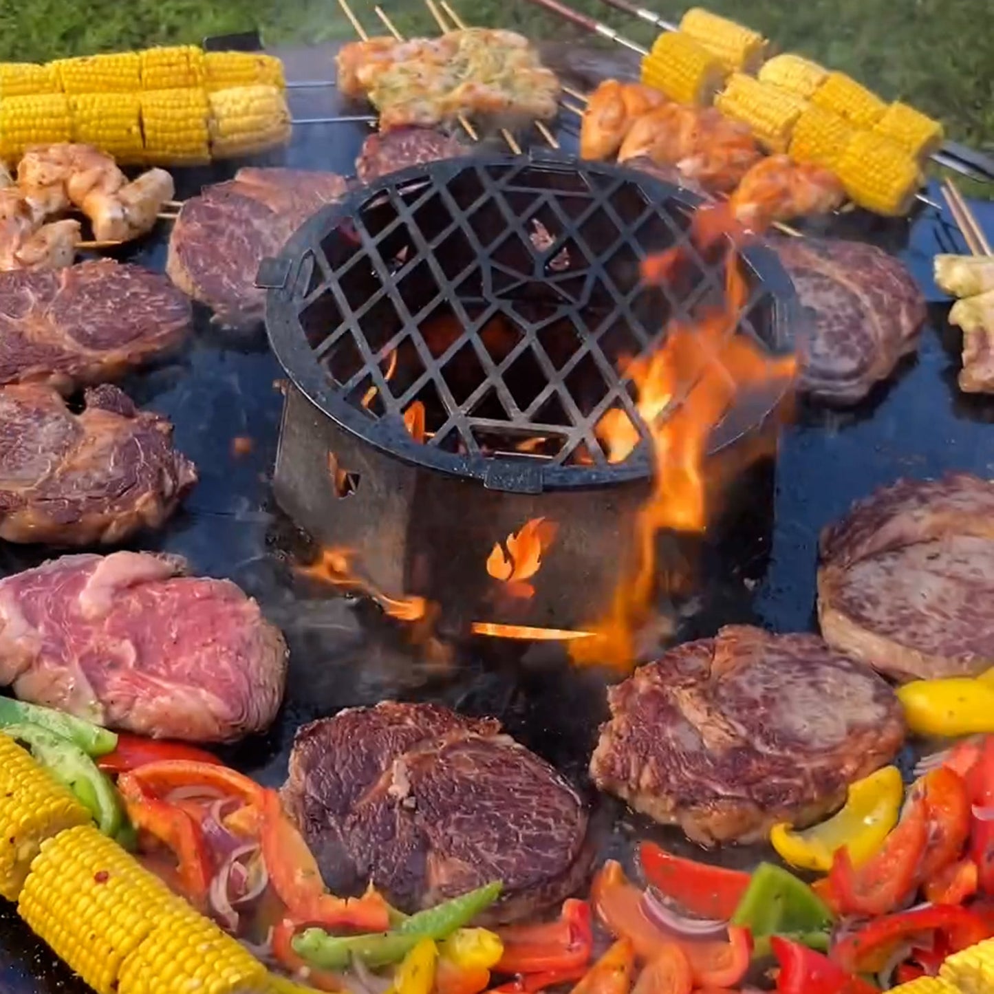 Szenebild der FENNEK Feuerplatte - 96 cm. Auf der Feuerplatte werden Steaks, Paprika, Mais und Spieße gegrillt.