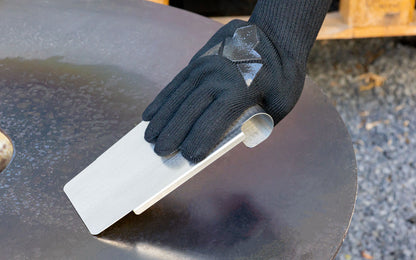 Grillspachtel wird von Handschuh gehalten und schabt auf Feuerplatte