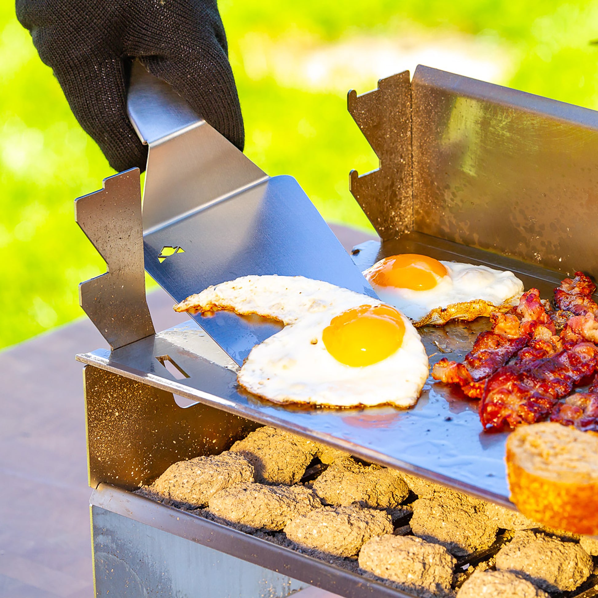 Schwarzer Grillhandschuh hebt mit Edelstahl BBQ-Wender ein Spiegelei vom Grill mit Plancha.