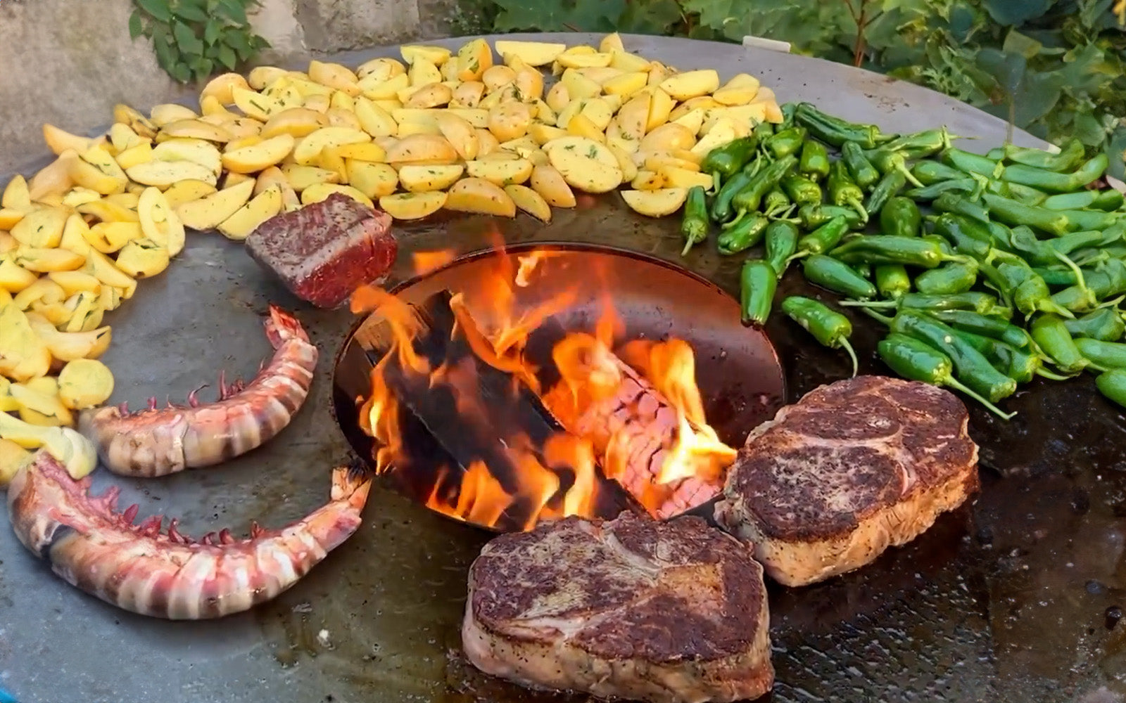 Leckere Schrimps, Steaks, Bratkartoffeln und Pimientos werden auf einer FENNEK Feuerplatte gegrillt. Im ø 30 cm Loch der Feuerplatte ragt ein loderndes Feuer hervor.