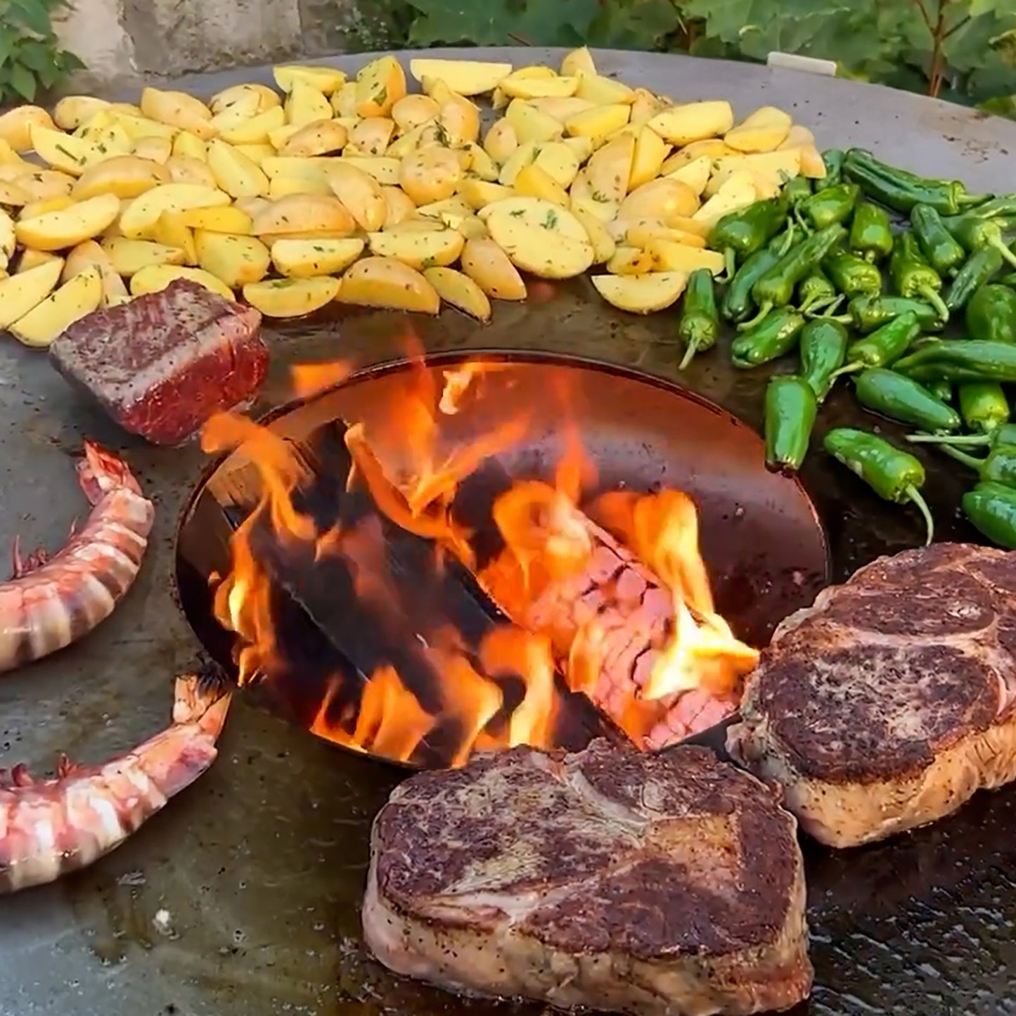 Brutzelnde Steaks, Shrimps, Bratkartoffeln und kleine Paprika auf der FENNEK Feuerplatte! Im mittleren Loch der Platte ist das Feuer erkennbar