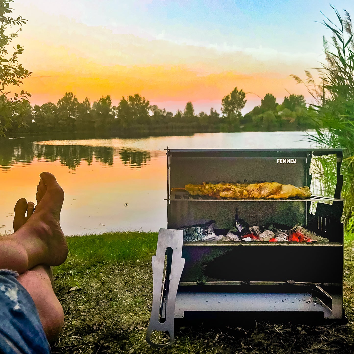 Fennek Grill mit glühender Kohle und Grillfleisch auf dem Rost bei Sonnenuntergang am See.