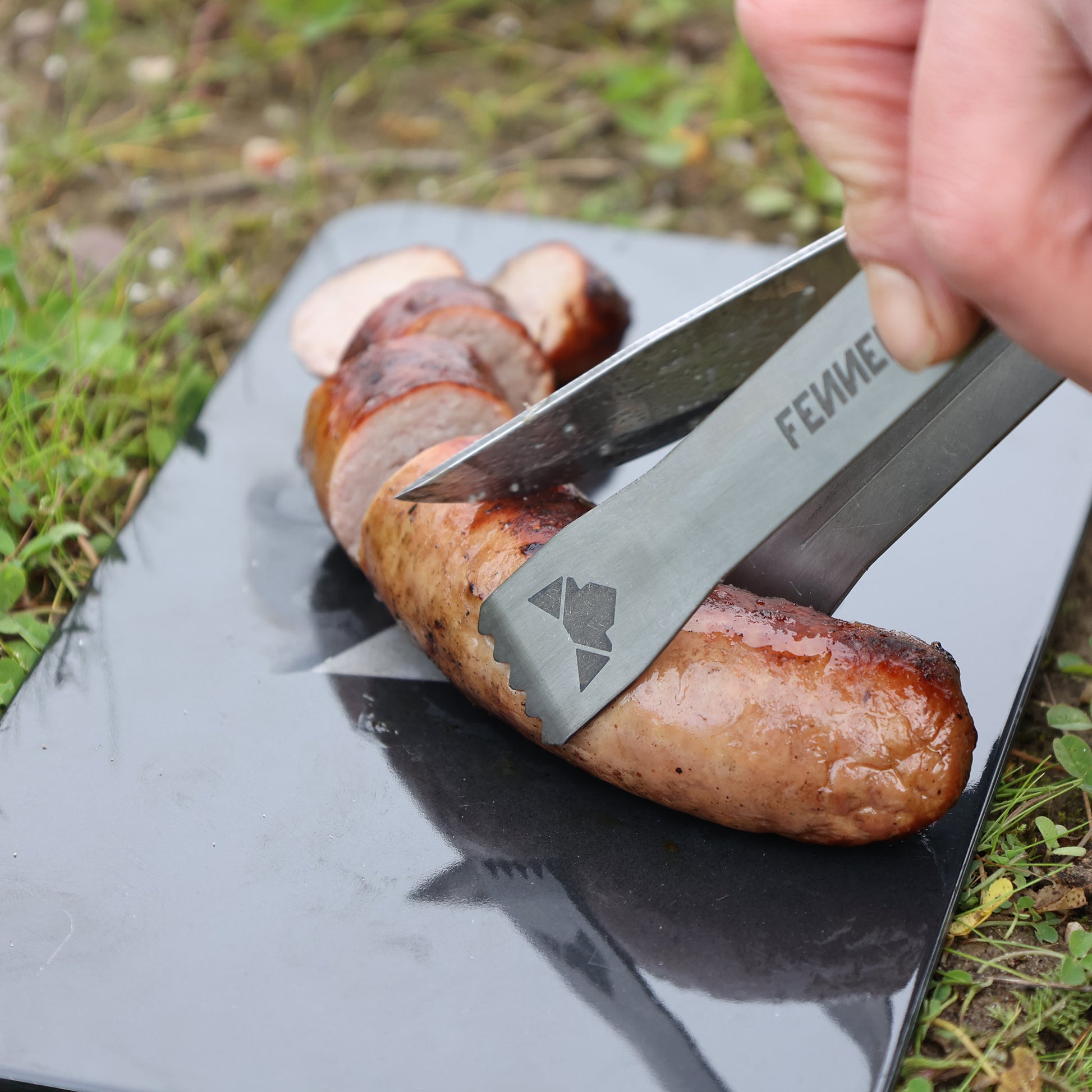 Kleine Grillzange aus Titan mit laser-graviertem FENNEK-Logo und Titanium-Schriftzug im Einsatz beim Schneiden einer Bratwurst
