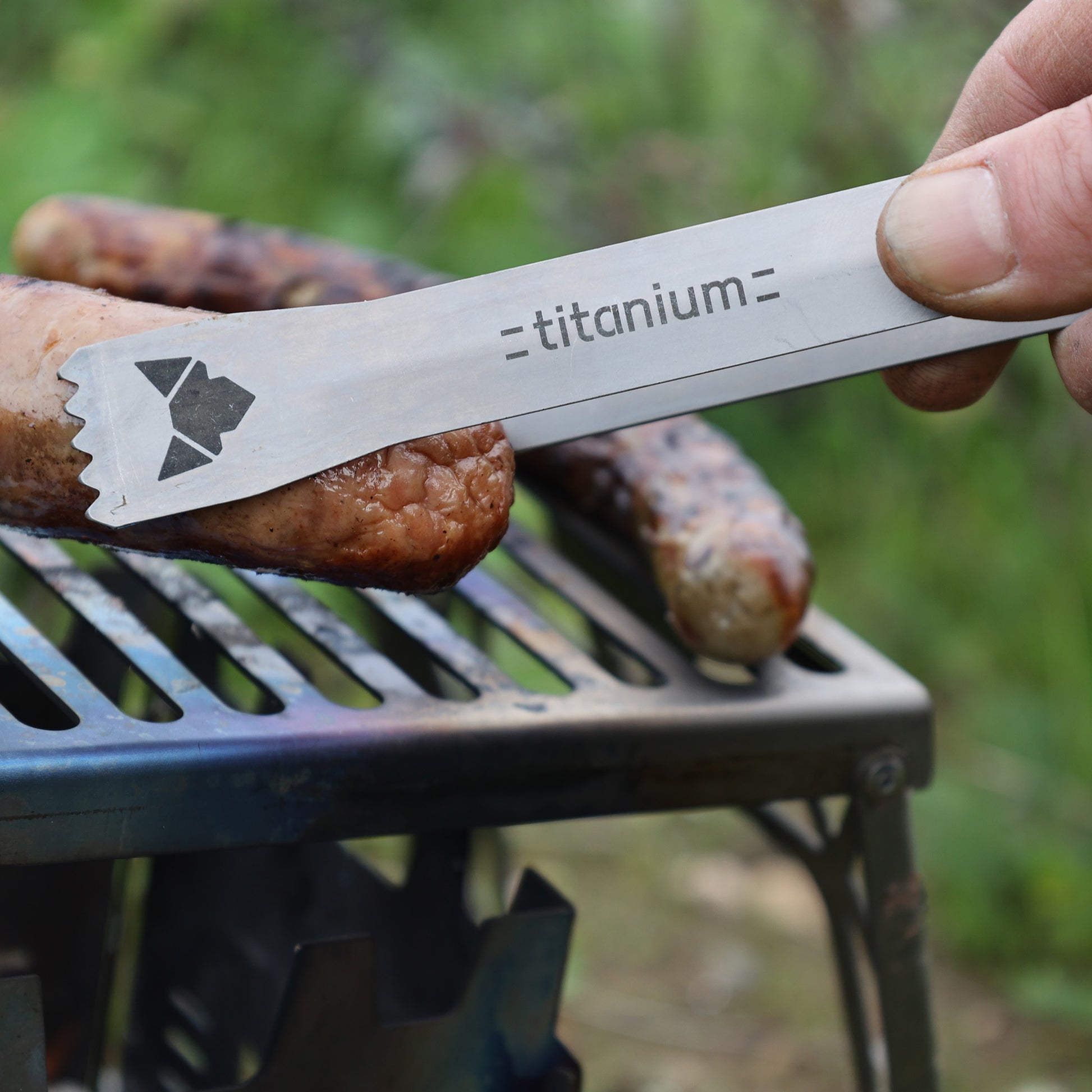 Kleine Grillzange aus Titan mit laser-graviertem FENNEK-Logo und Titanium-Schriftzug im Einsatz beim Greifen einer Bratwurst