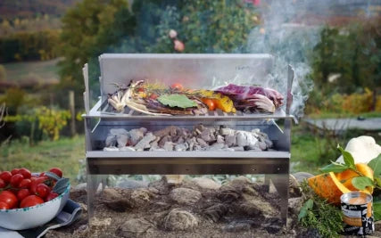 Ein FENNEK Grill im Garten. Darauf wird Fleisch und Gemüse gebraten. Links eine Schale mit Tomaten.
