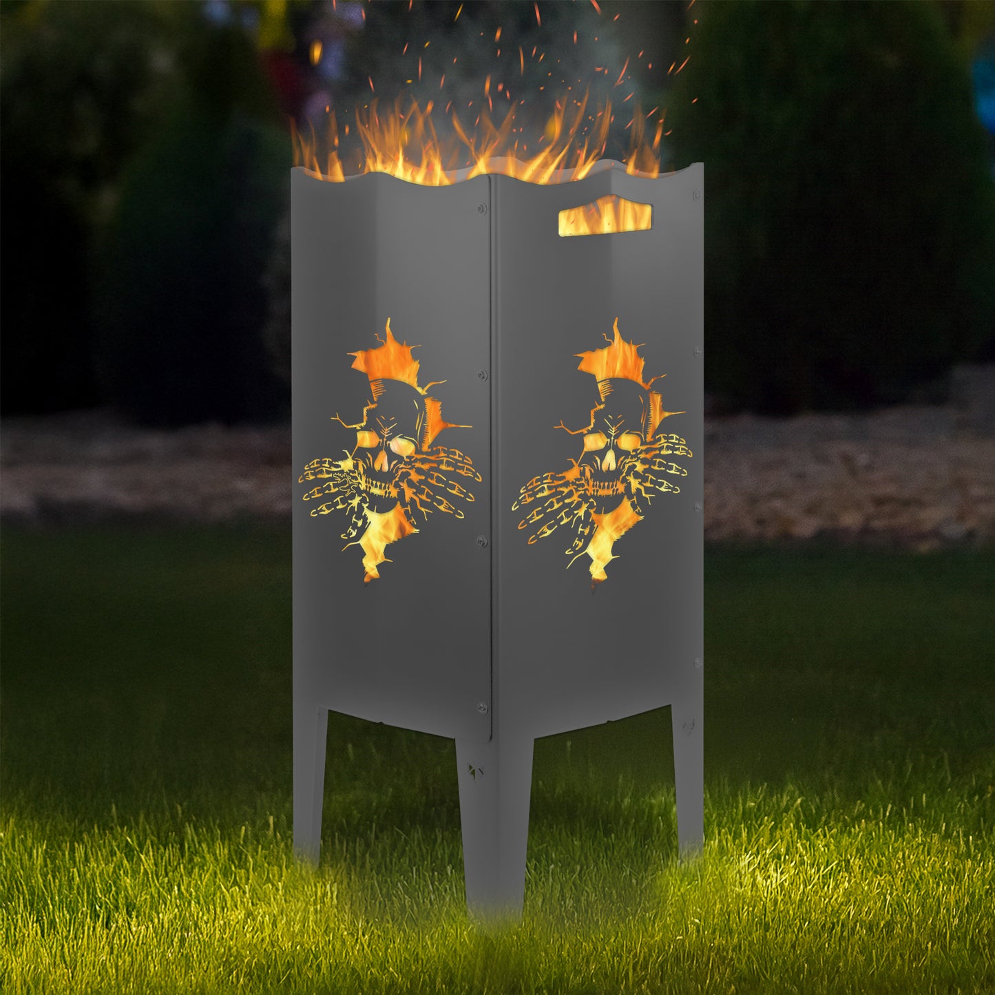 FENNEK Feuersäule Stahl mit dem Motiv Totenkopf - in der Feuersäule brennt ein Feuer, man sieht das Motiv sehr schön leuchten in der Abenddämmerung in einem Garten. Die Feuersäule steht auf Gras.