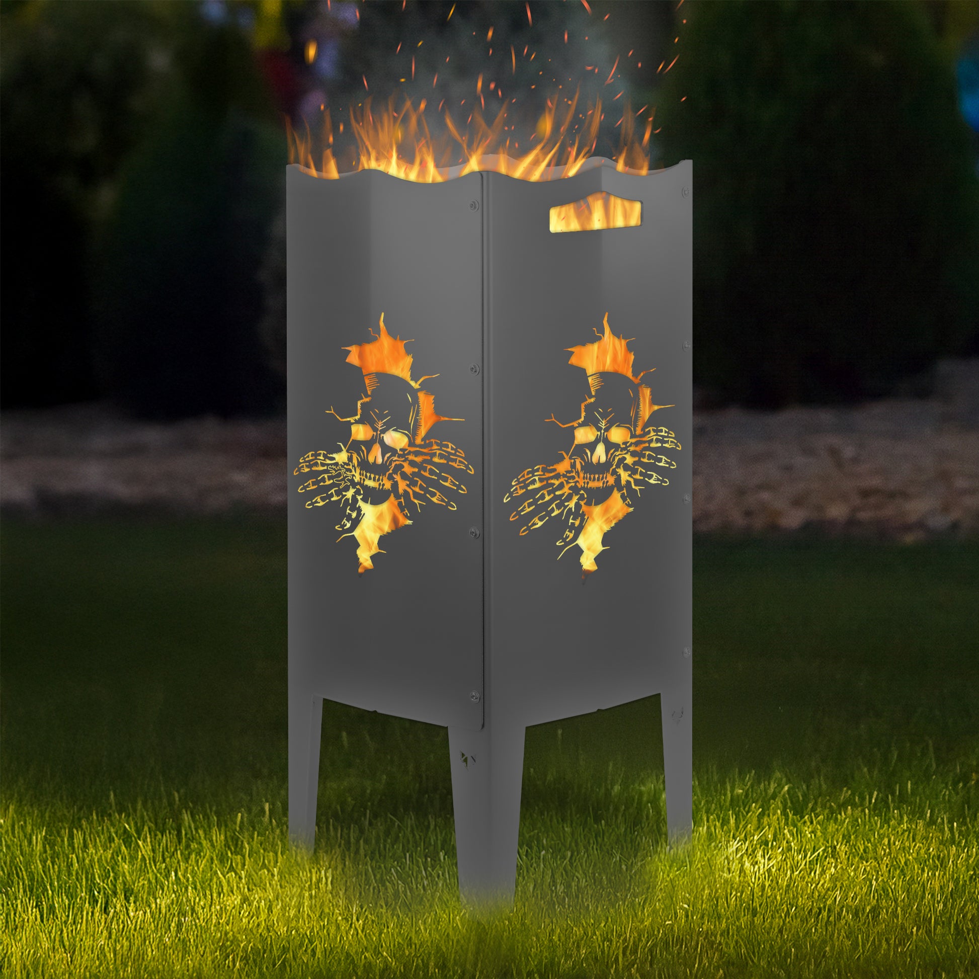 FENNEK Feuersäule Stahl mit dem Motiv Totenkopf - in der Feuersäule brennt ein Feuer, man sieht das Motiv sehr schön leuchten in der Abenddämmerung in einem Garten. Die Feuersäule steht auf Gras.