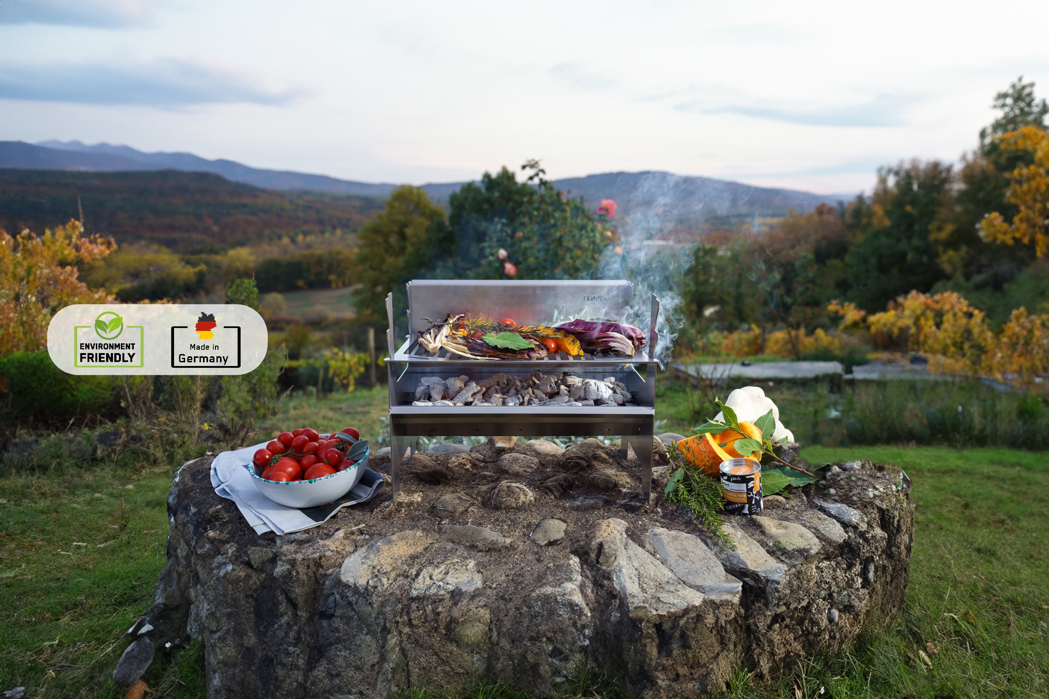 Der FENNEK 2.0 Holzkohlegrill steht auf einem Steinvorsprung im Grünen. Die Kohle glüht und auf dem Rost liegen saftige Steaks und Gemüse.