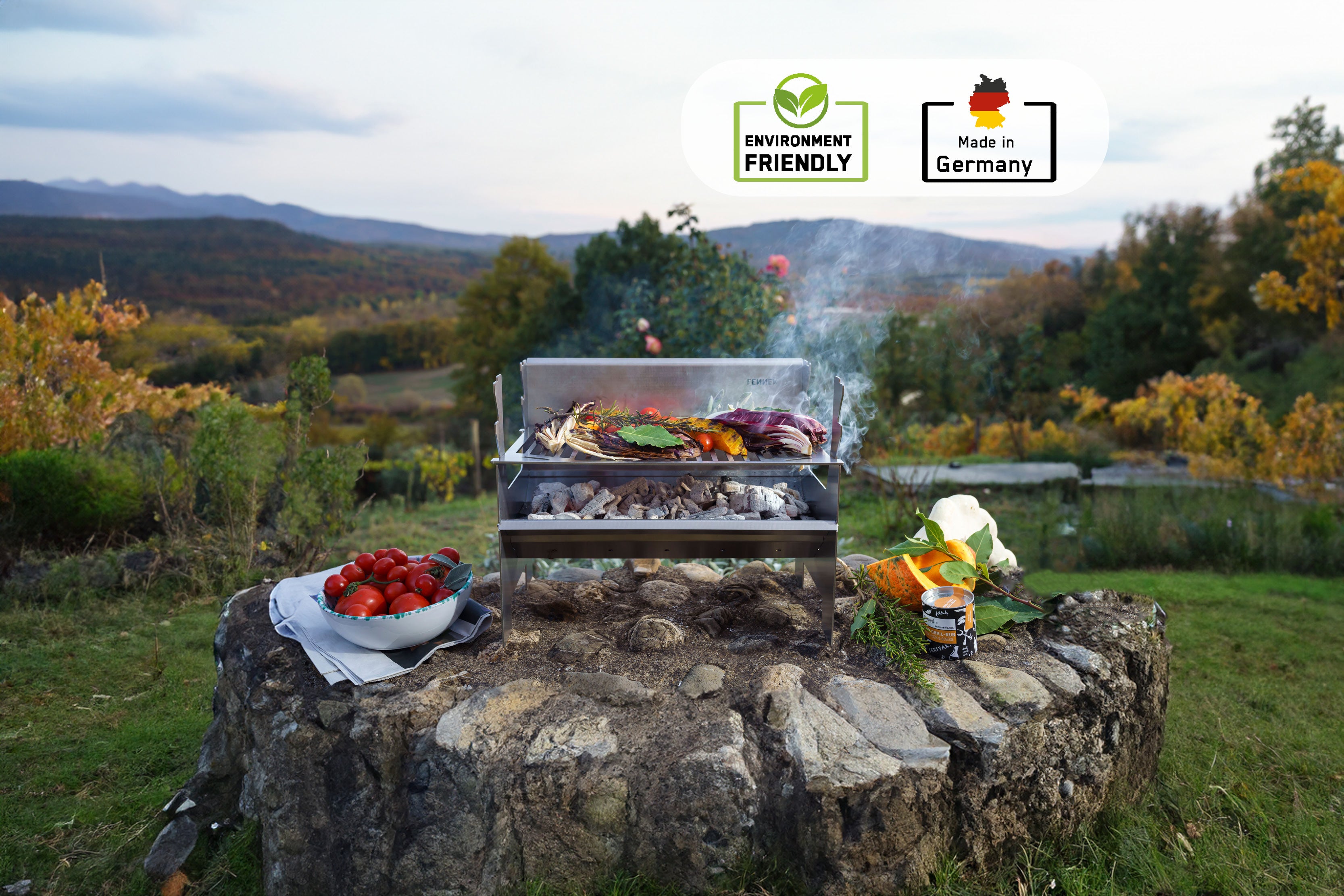 Der FENNEK 2.0 Holzkohlegrill steht auf einem Steinvorsprung im Grünen. Die Kohle glüht und auf dem Rost liegen saftige Steaks und Gemüse.