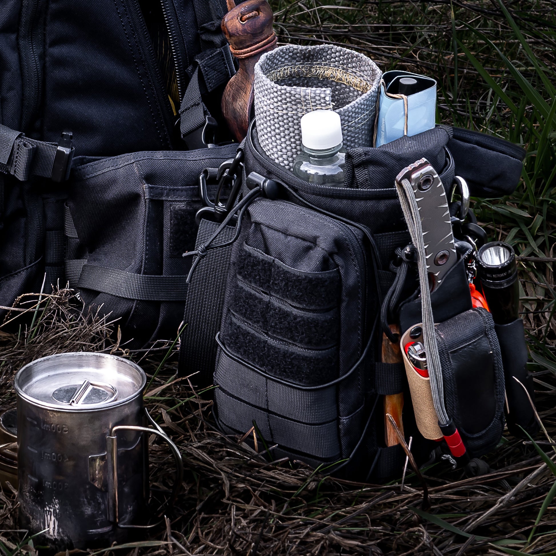 Die Zubehörtasche steht neben dem Backpack One2explore auf einer Wiese. Der Deckel der Zubehörtasche ist geöffnet und zeigt den sich darin befindenden Inhalt. Zu erkennen sind unter anderem die FENNEK Flammschutzmatte, ein Messer, ein Feuerzeug und eine Taschenlampe.