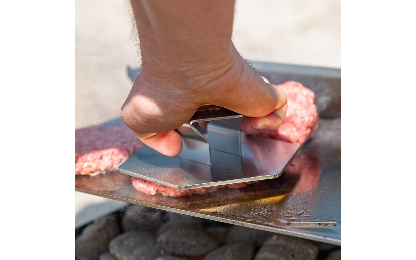 Eine Hand hält 6-eckige Hamburger Presse aus Edelstahl und presst einen Burger Patty auf der Plancha eines Holzkohlegrills.