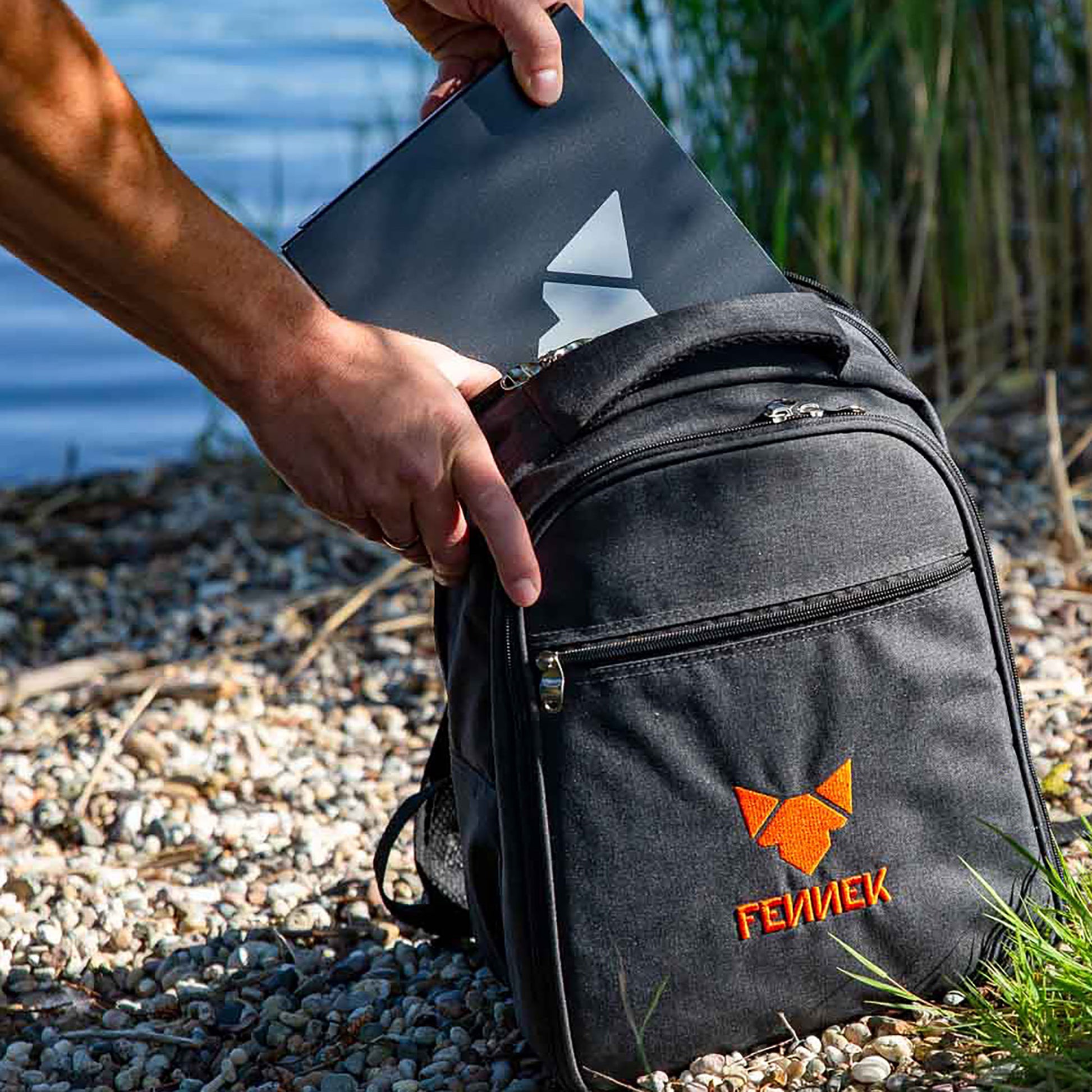 Der FENNEK Grill wird am Kiesstrand aus einem dunkelgrauem Rucksack mit FENNEK Logo-Stickerei gezogen und unscharf im Hintergrund ein See und Schilf.