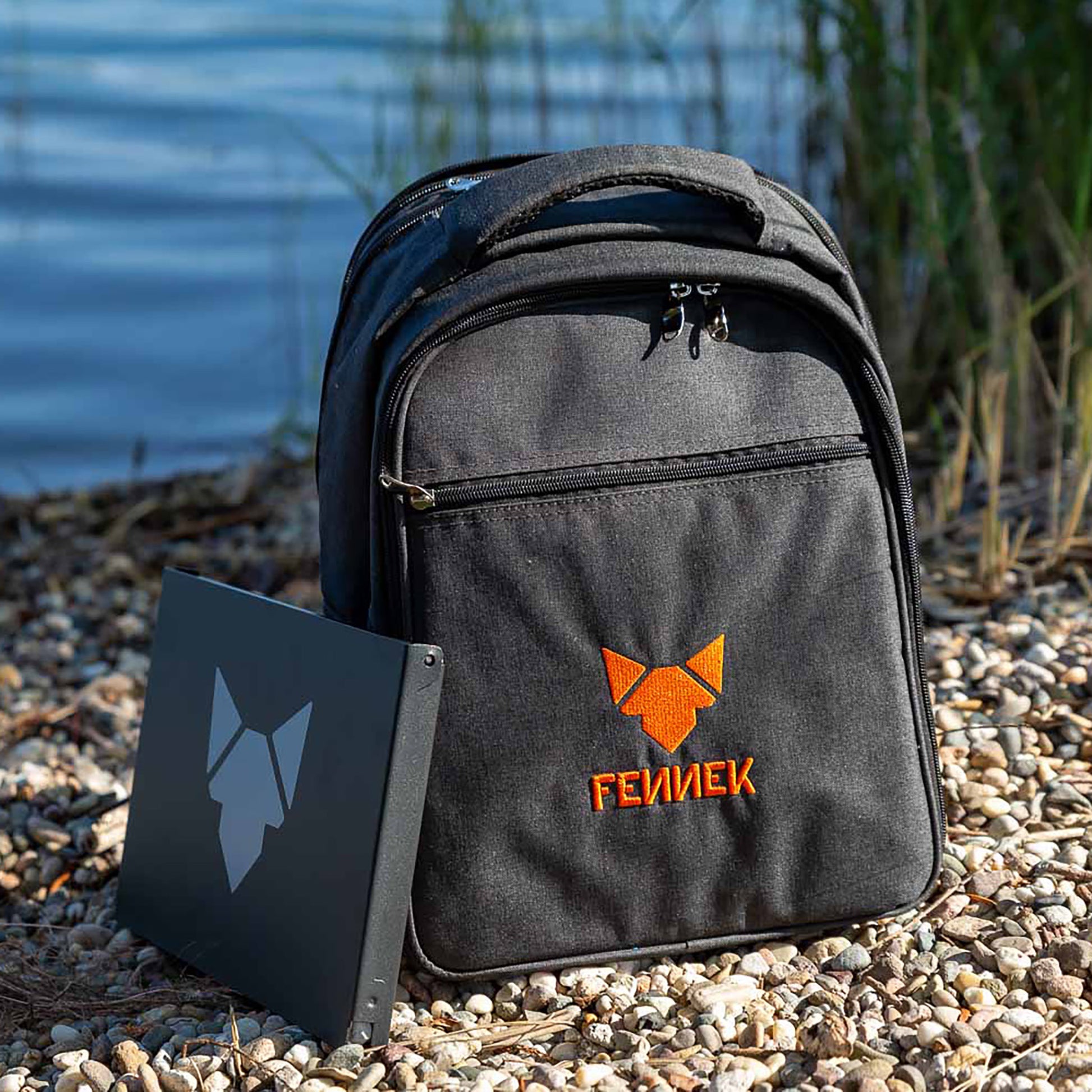 Dunkelgrauer Rucksack mit FENNEK Logo-Stickerei auf der Front und angelehnt der FENNEK Grill in einer schwarzen Alubox mit hellgrauem FENNEK Logo-Aufdruck am Kiesstrand und unscharf im Hintergrund ein See und Schilf.
