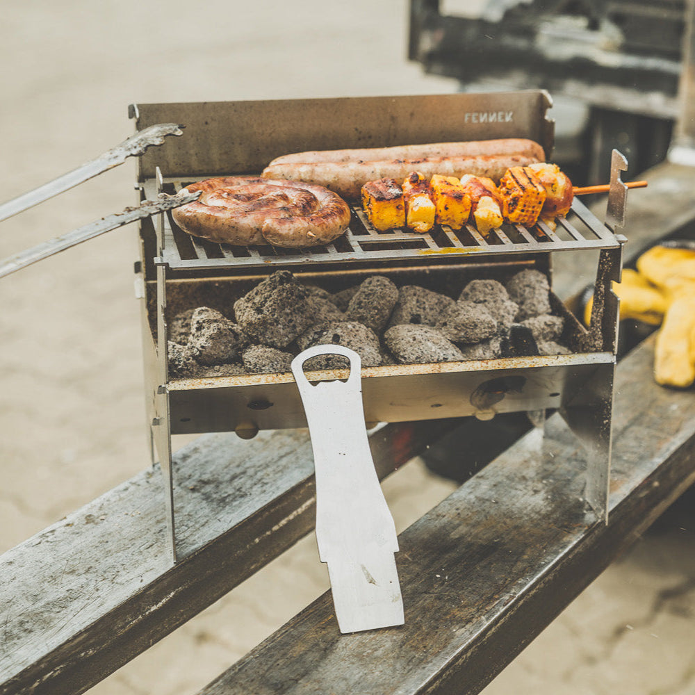 FENNEK Light Grill in Szene auf einer Gabelstaplergabel in Szene gesetzt. Das darauf brutzelnde Fleisch wird mit der Grillzange gewendet. Am FENNEK Light angelehnt sieht man den Rostheber/Flaschenöffner.