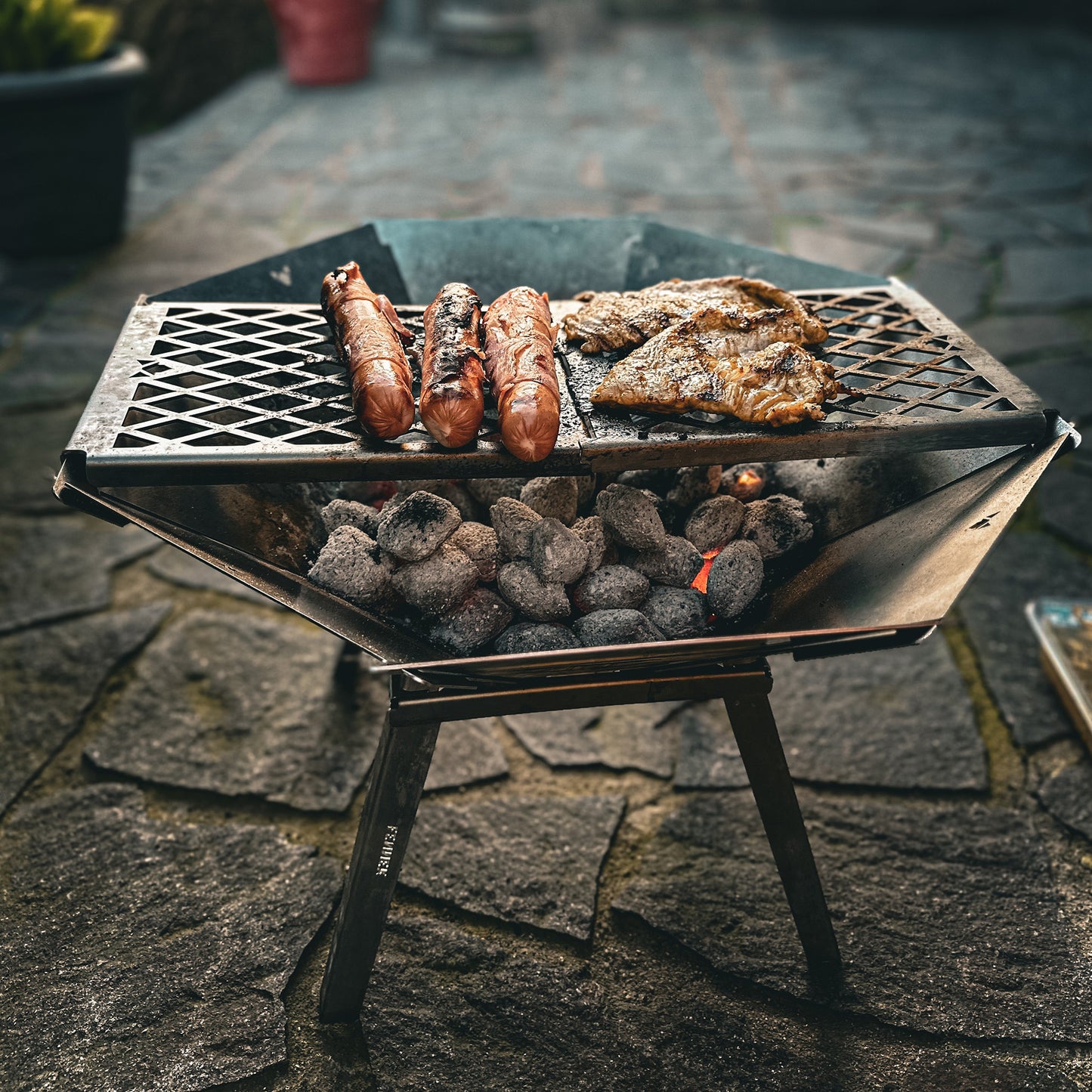Die mobile, kompakte, steckbare Feuerschale Oktagon aus Edelstahl steht aufgebaut mit Füßen und Grillrost auf einer Steinterrasse. Es werden Berner Würstchen und Hähnchensteaks über glühender Glut gegrillt.