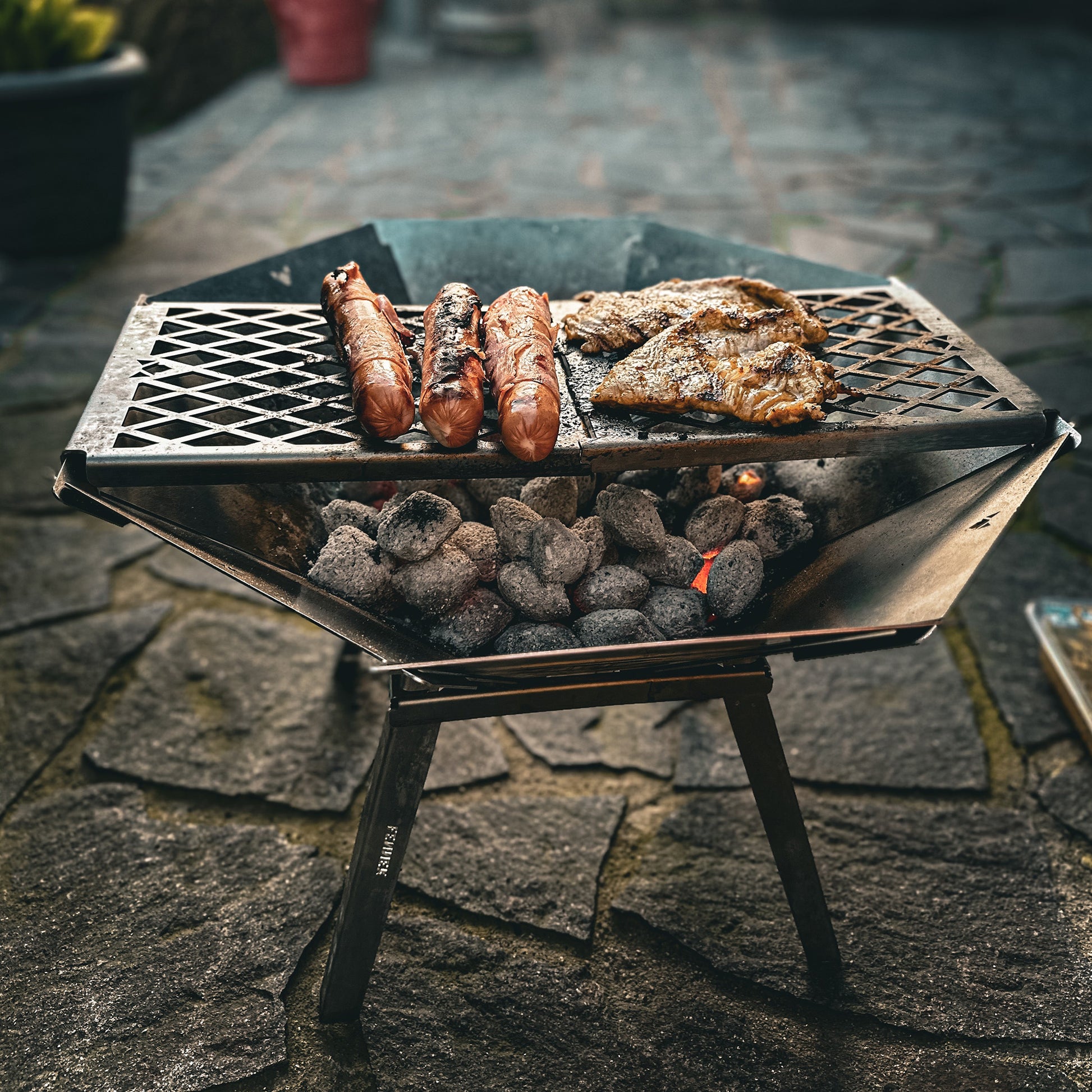 Die mobile, kompakte, steckbare Feuerschale Oktagon aus Edelstahl steht aufgebaut mit Füßen und Grillrost auf einer Steinterrasse. Es werden Berner Würstchen und Hähnchensteaks über glühender Glut gegrillt.